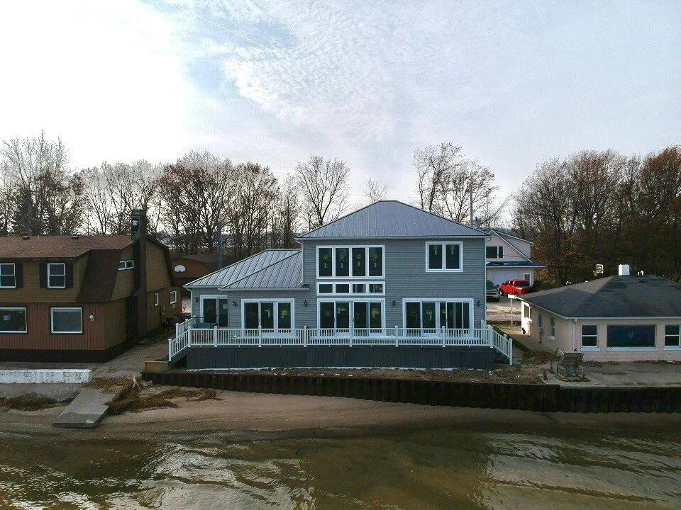 Gray waterfront house with large windows and a metal roof beside smaller homes under a cloudy sky