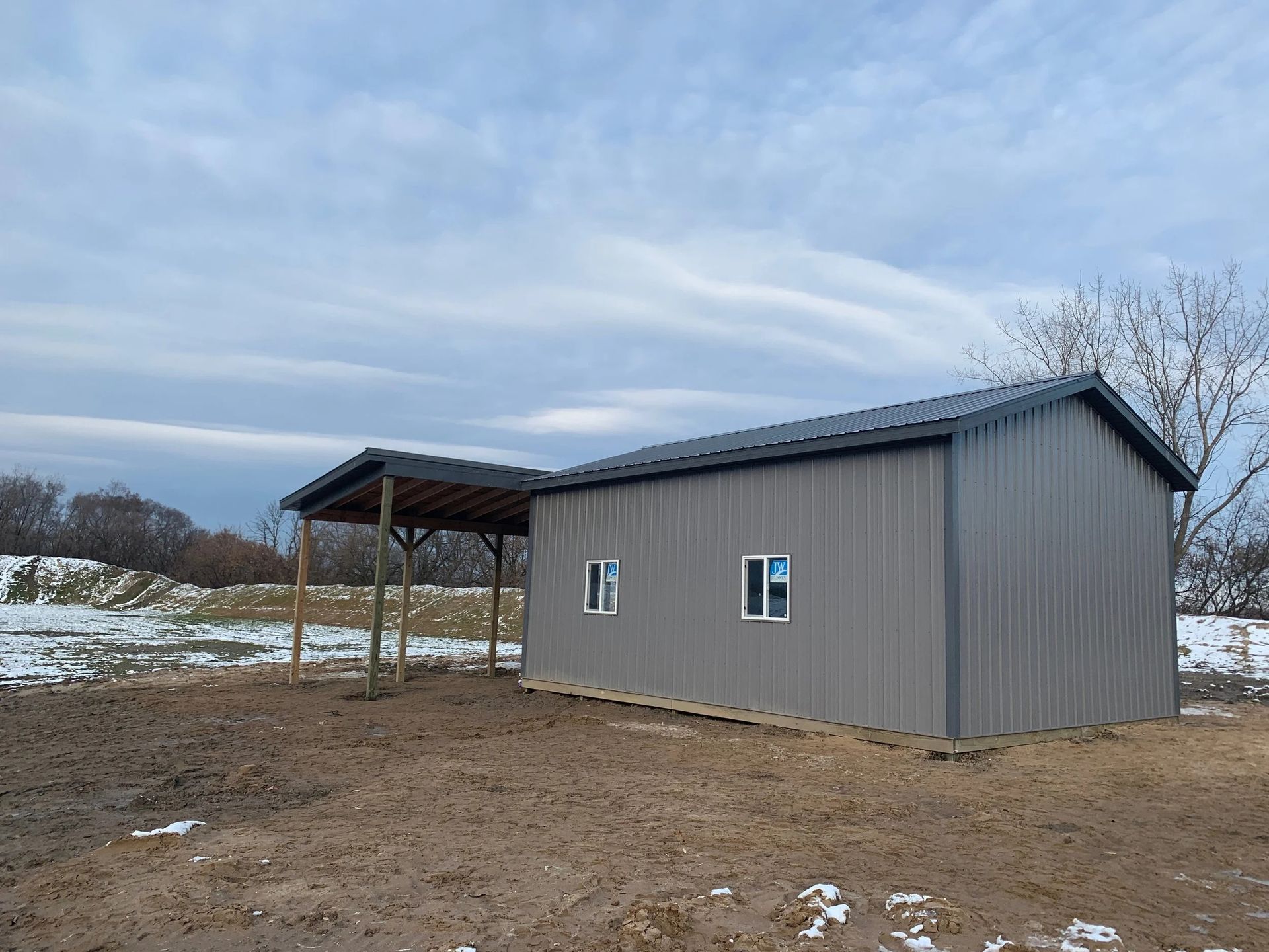 Gray metal shed with a covered porch in a snowy rural field under a cloudy sky
