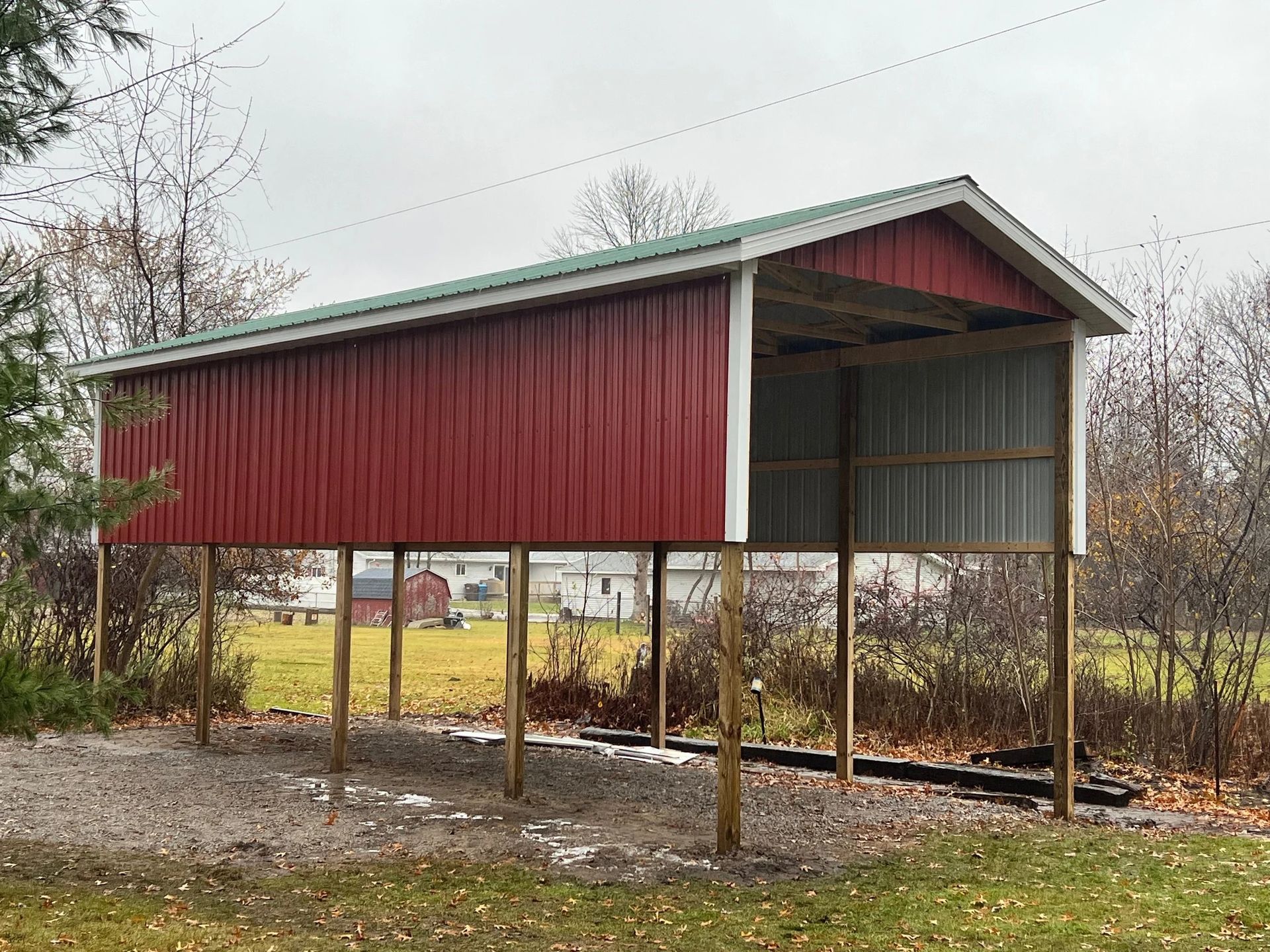 Red metal shed on posts in a bare rural yard, with trees and a cloudy sky behind it.