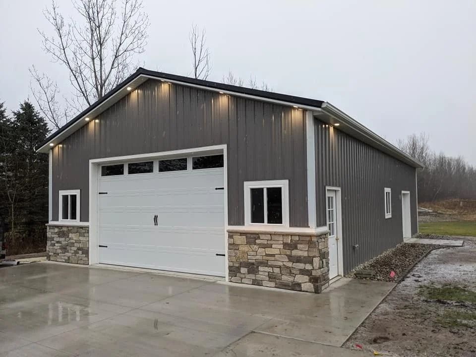 Gray metal garage with white door and stone base on a wet driveway under overcast skies