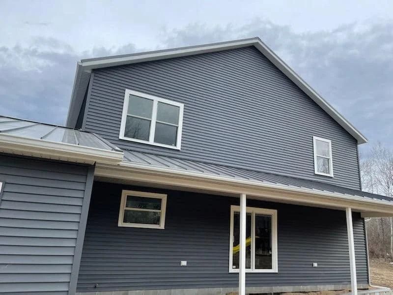 Blue-gray house exterior with white trim, covered porch, and dormer windows under a cloudy sky
