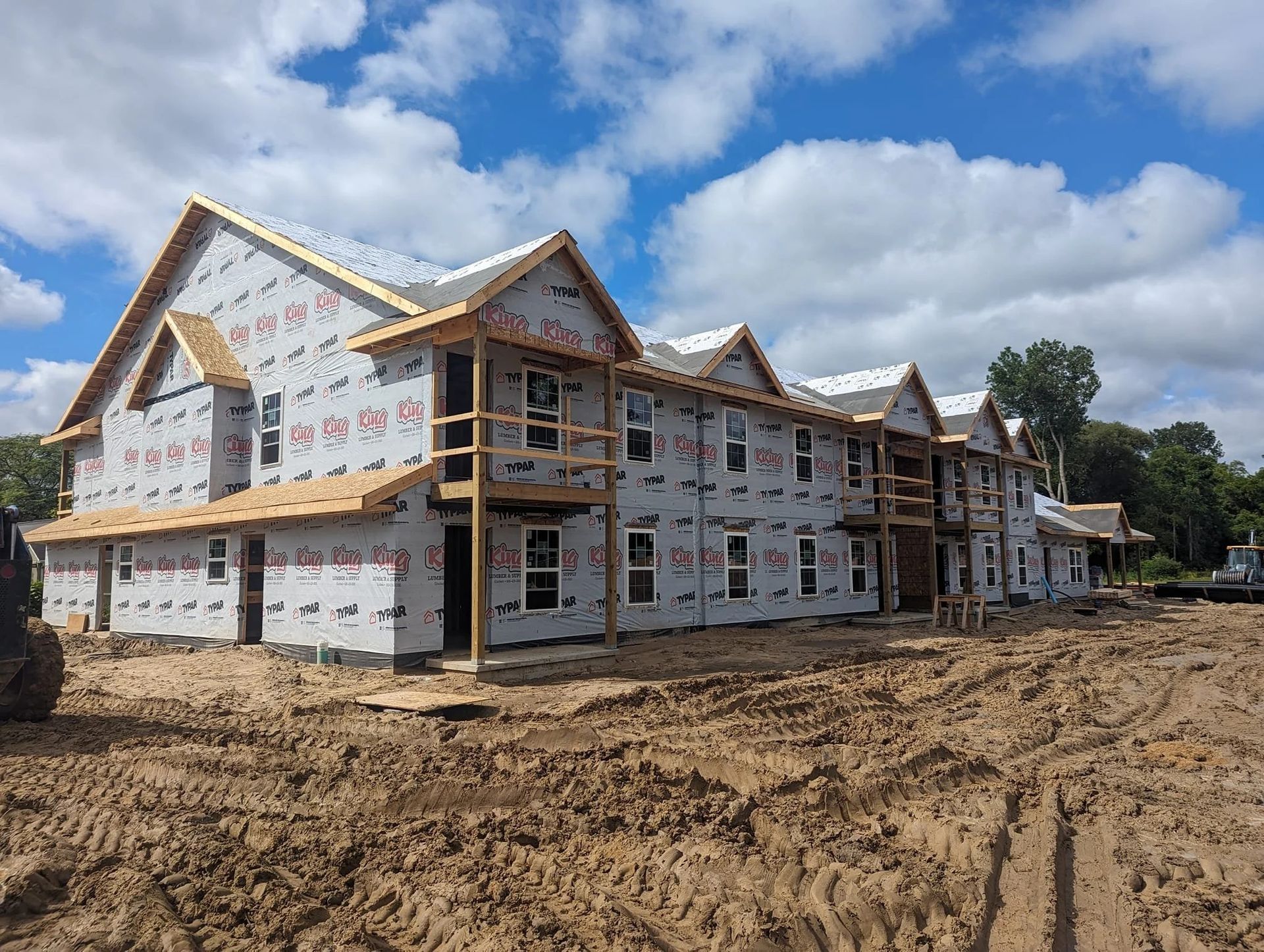 Large white house under construction on a muddy lot, with blue sky and clouds overhead.