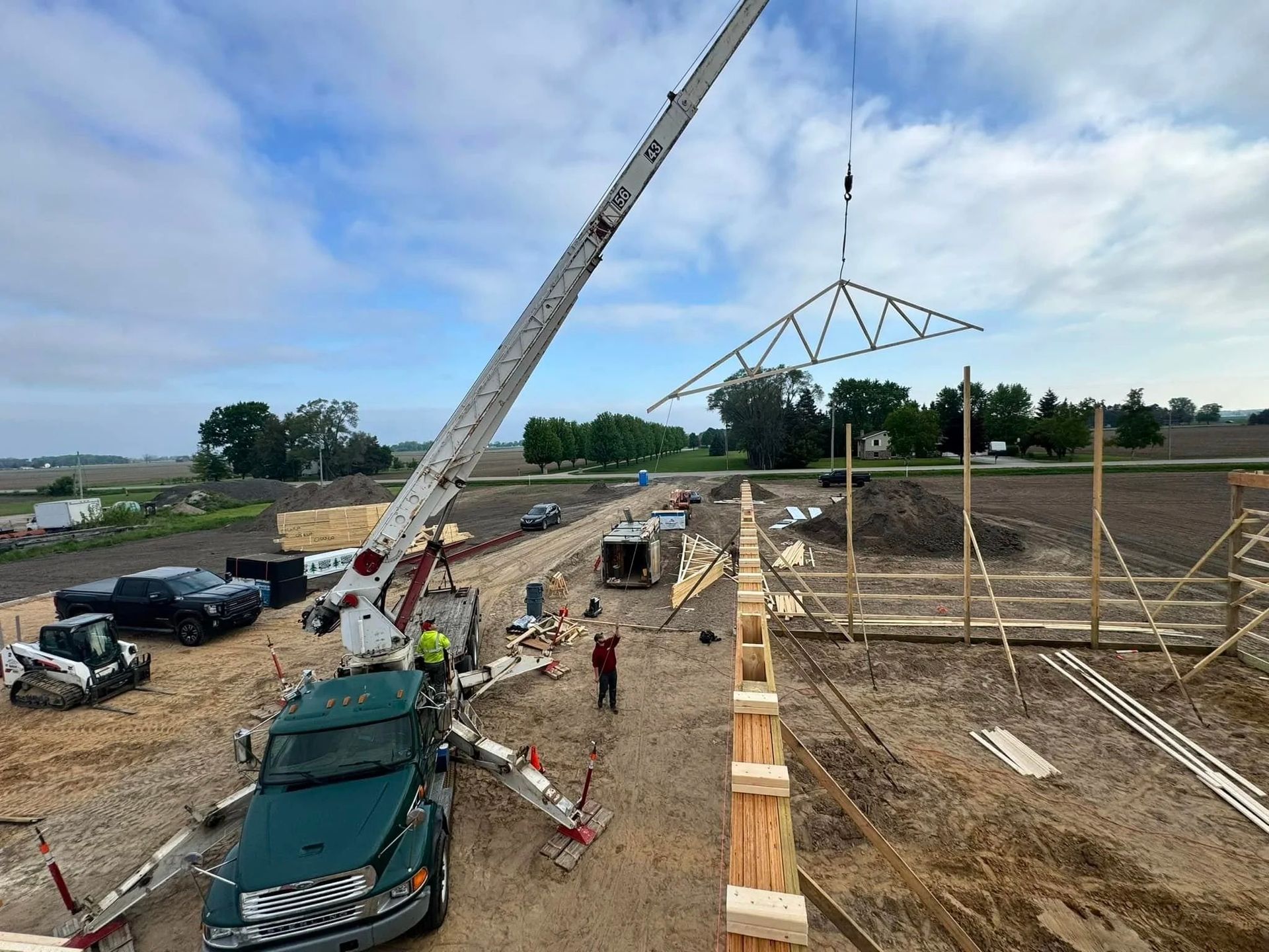 Crane lifting a roof truss at a construction site with vehicles and workers nearby