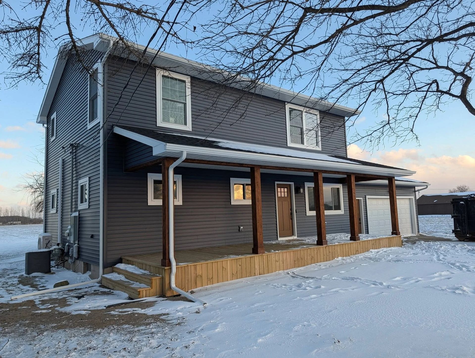 Snow-covered two-story gray house with a covered porch and bare trees in winter light