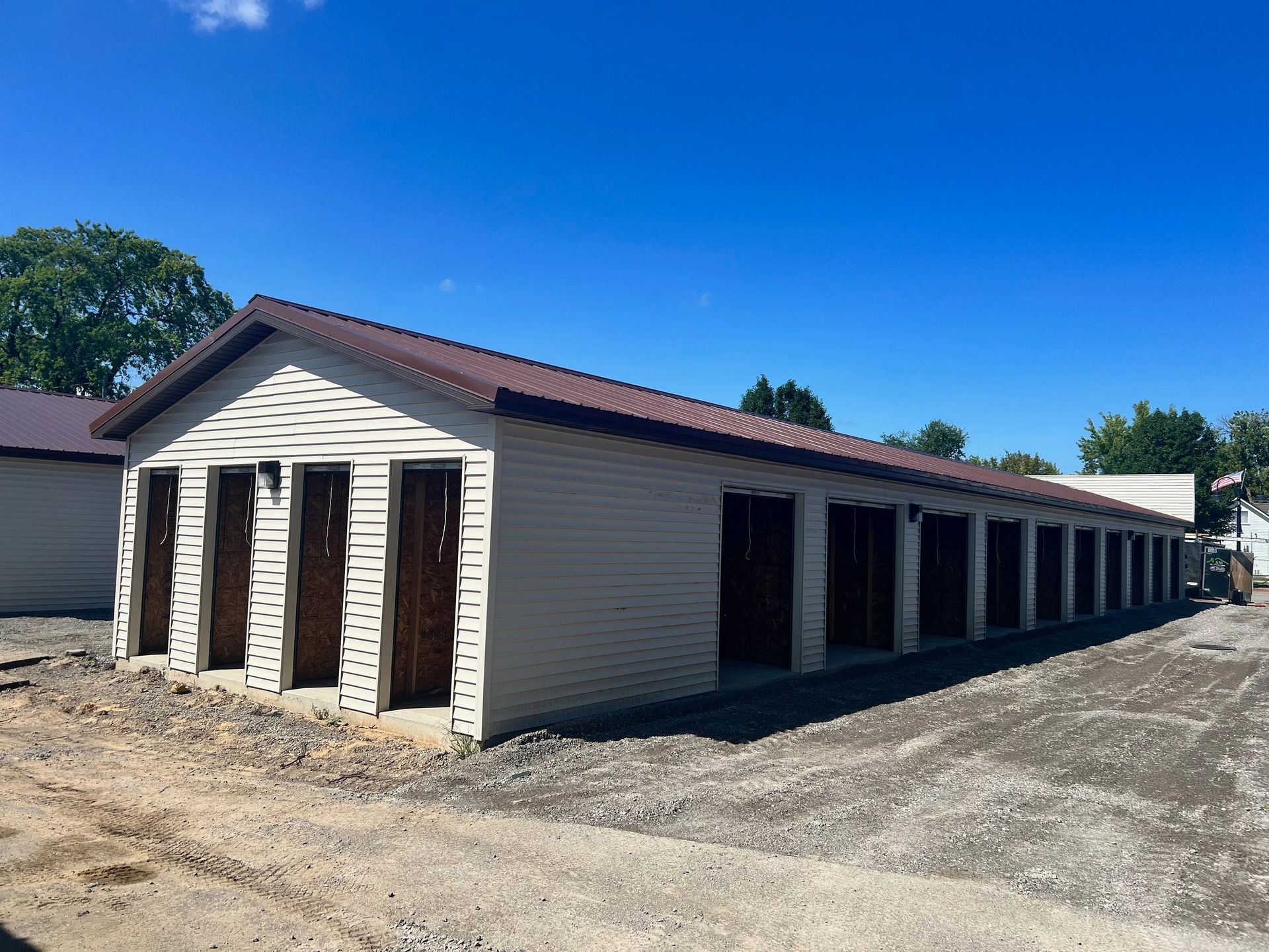 Row of beige storage units with dark brown doors under a blue sky, viewed from a dirt lot