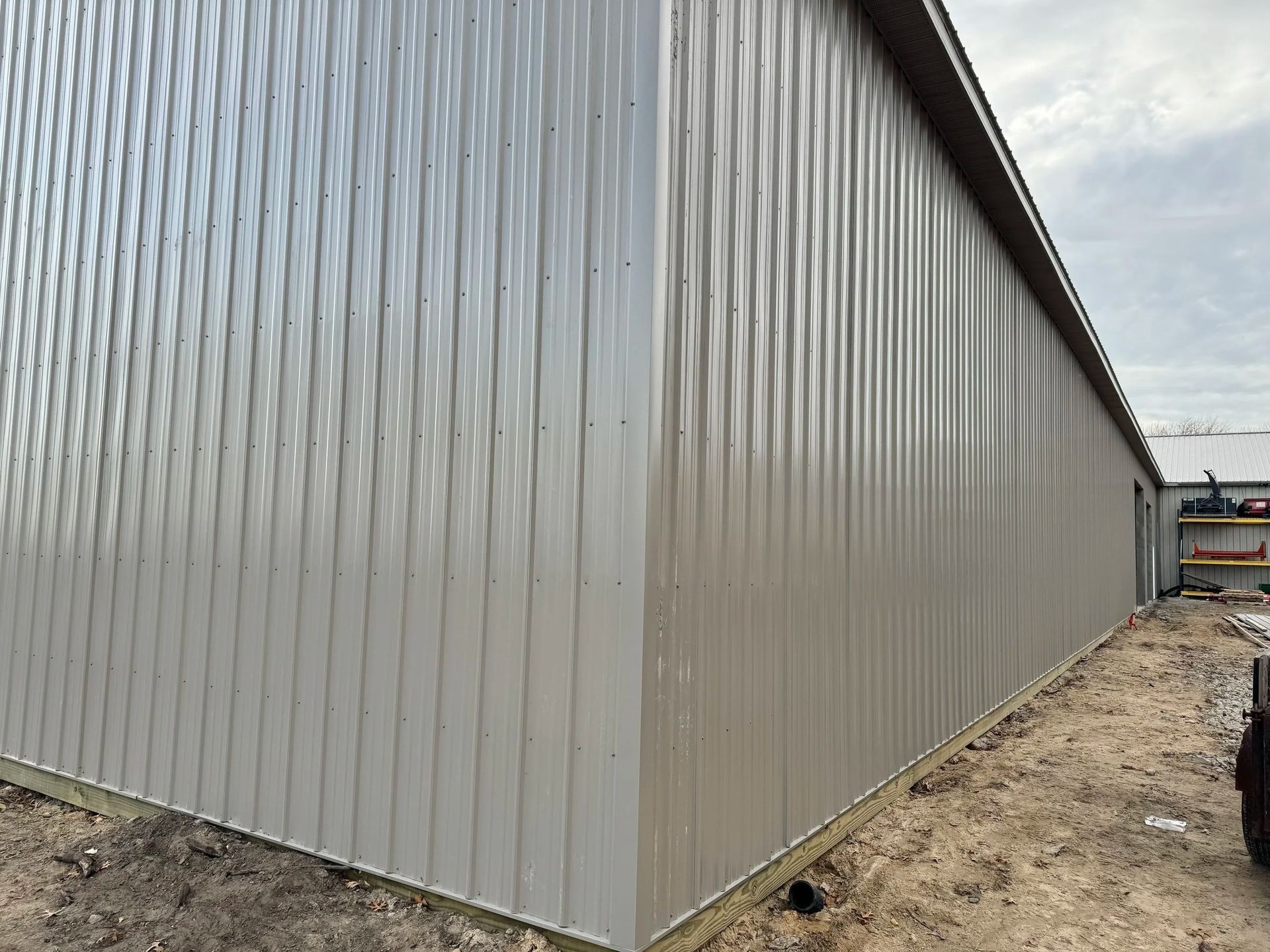 Large gray metal-sided building viewed from the corner on a dirt lot under a cloudy sky.