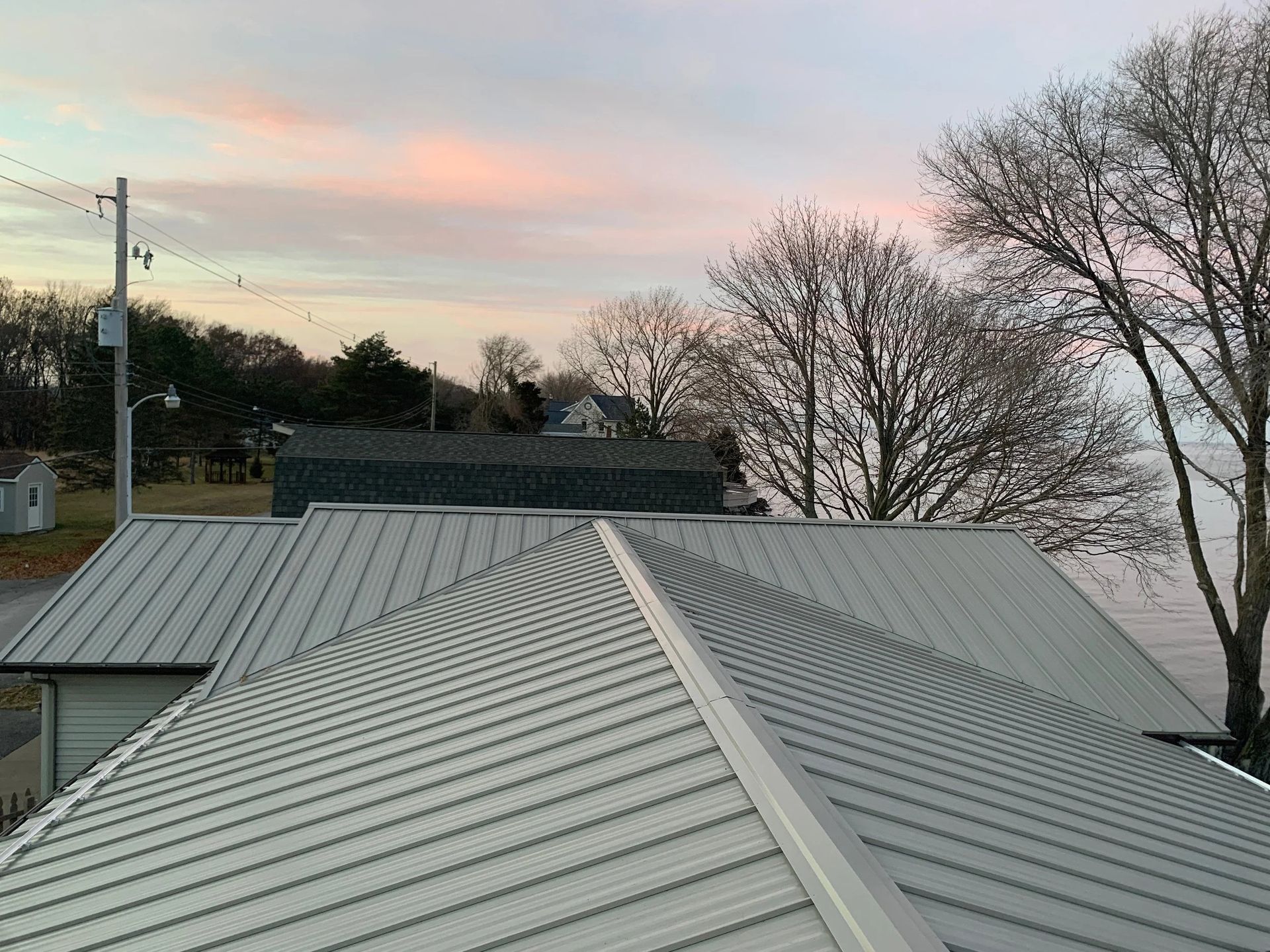 Rooftop with light gray metal panels at sunset, with trees and utility poles in the background