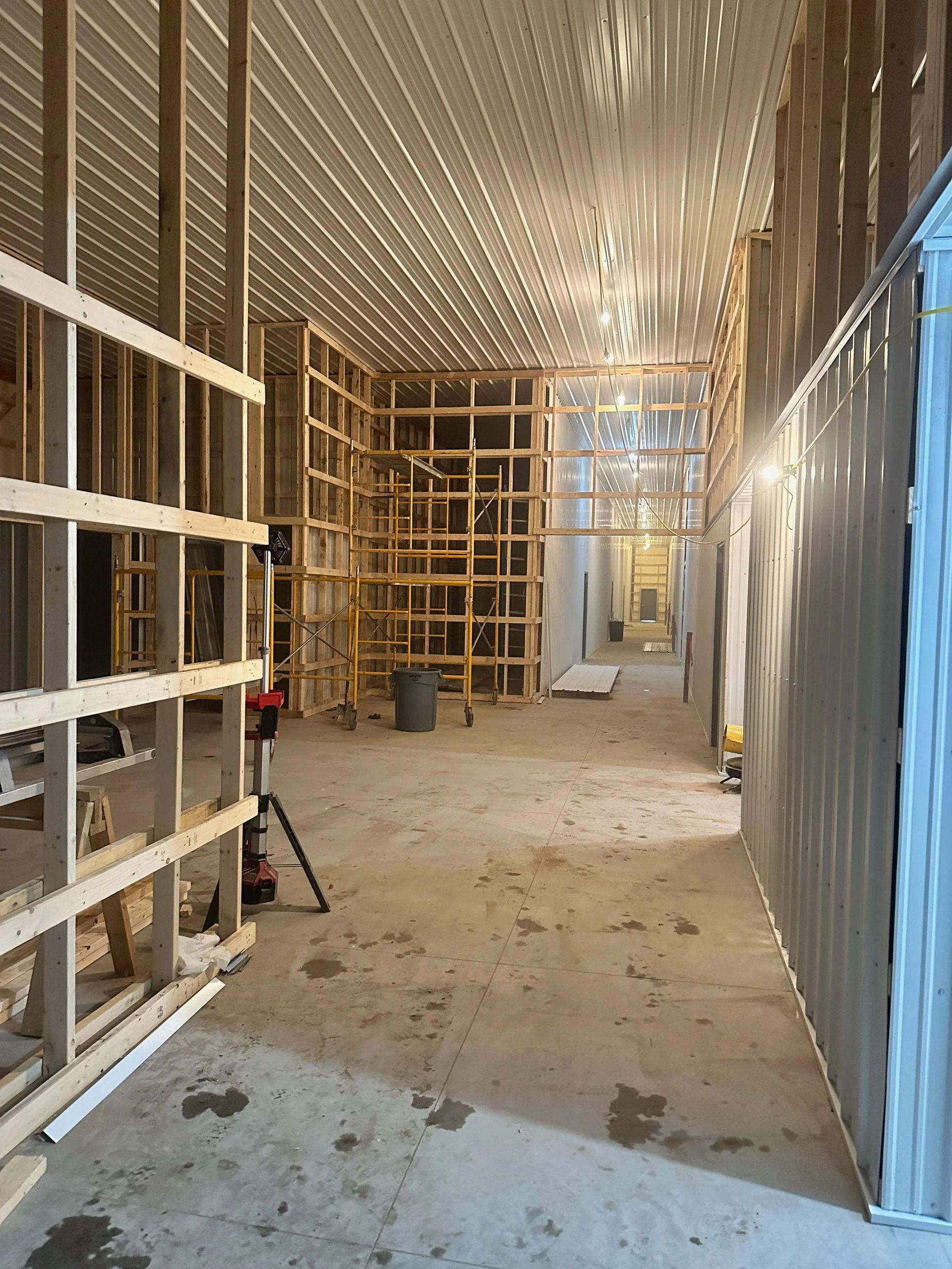 Narrow industrial hallway under construction with exposed framing, unfinished walls, and scattered debris on the floor.
