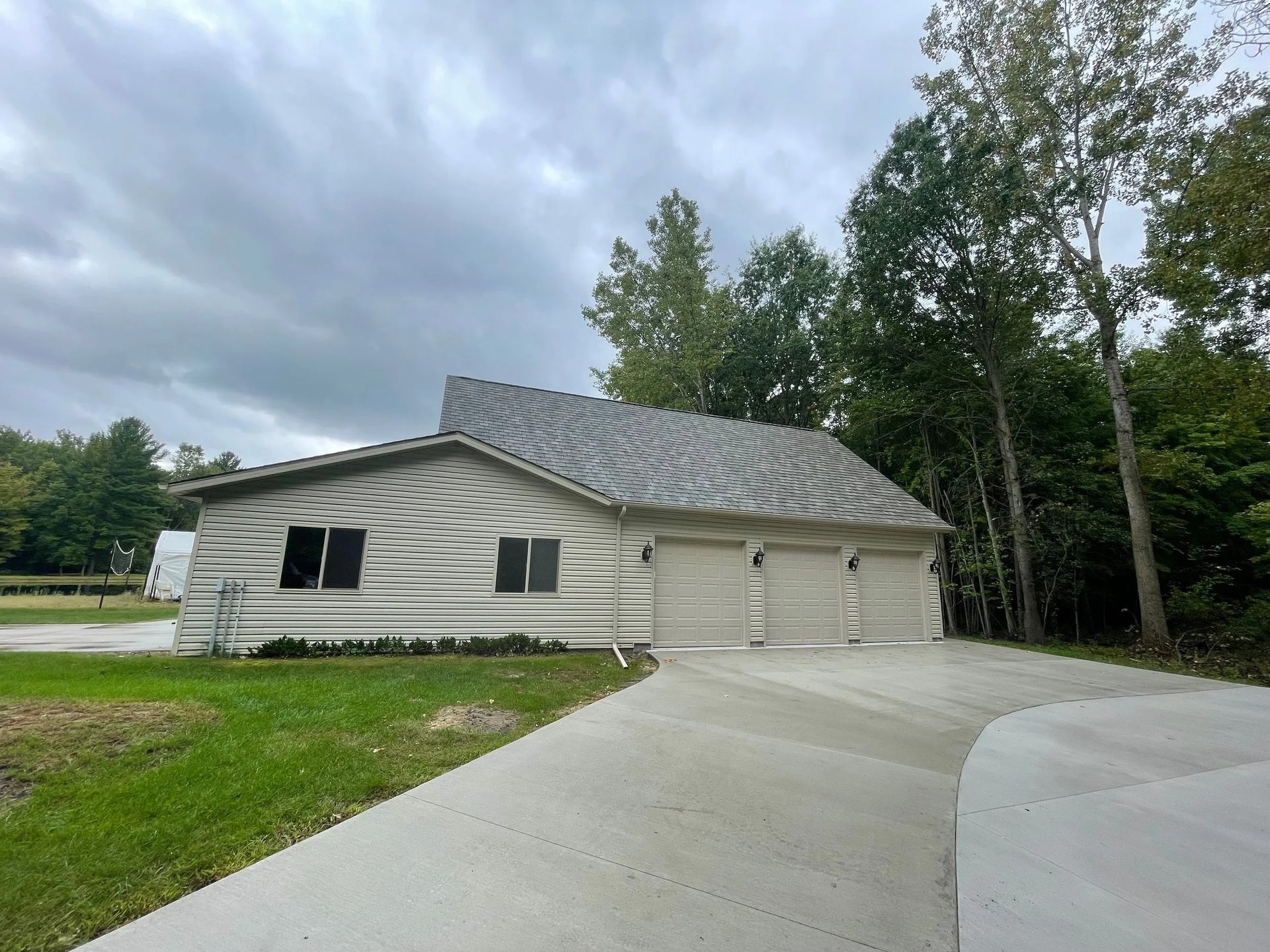 Single-story house with a curved driveway, attached garage, and cloudy sky in a wooded suburban setting