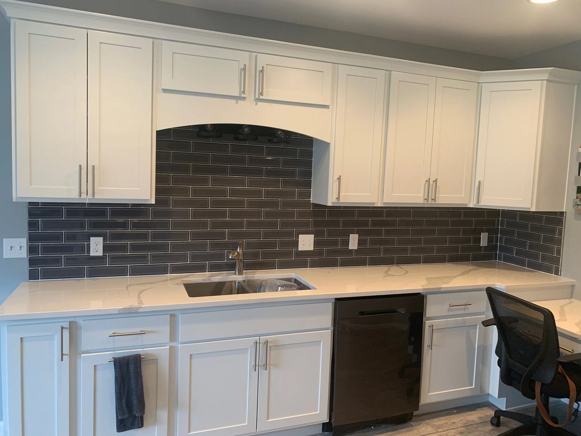 Modern kitchen with white cabinets, dark tile backsplash, light countertop, sink, and black dishwasher.