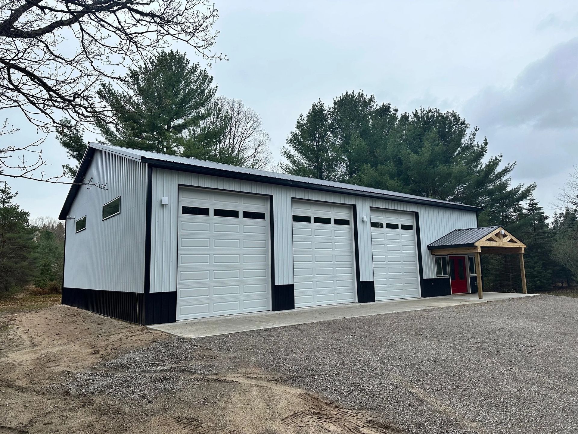 Gray metal garage with three white doors on a gravel driveway under cloudy skies