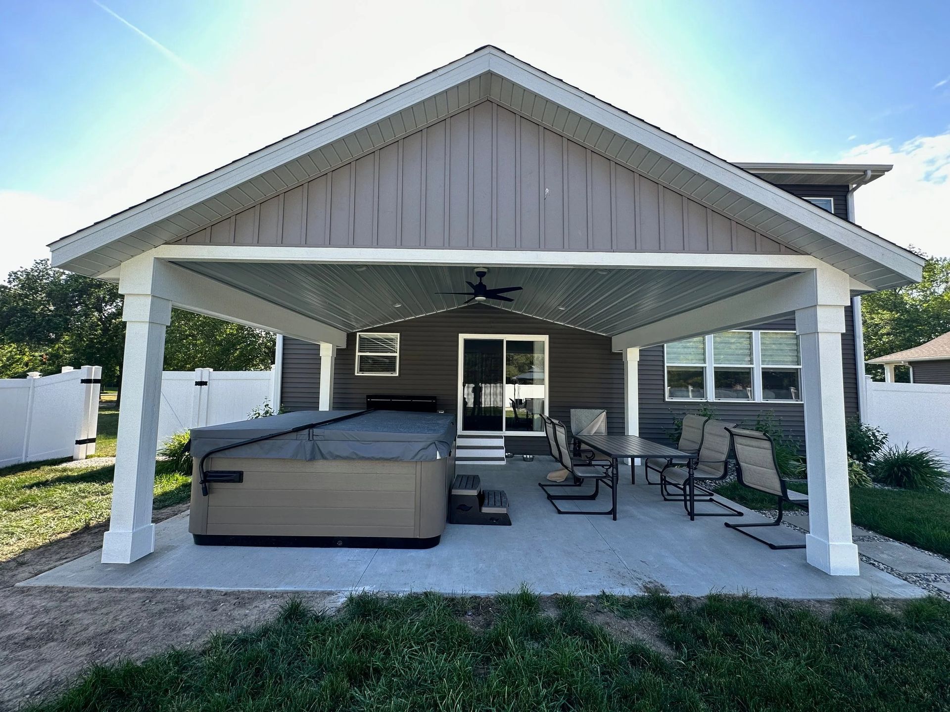 Covered patio with white posts, outdoor sofa and chairs, and a ceiling fan in front of a house.