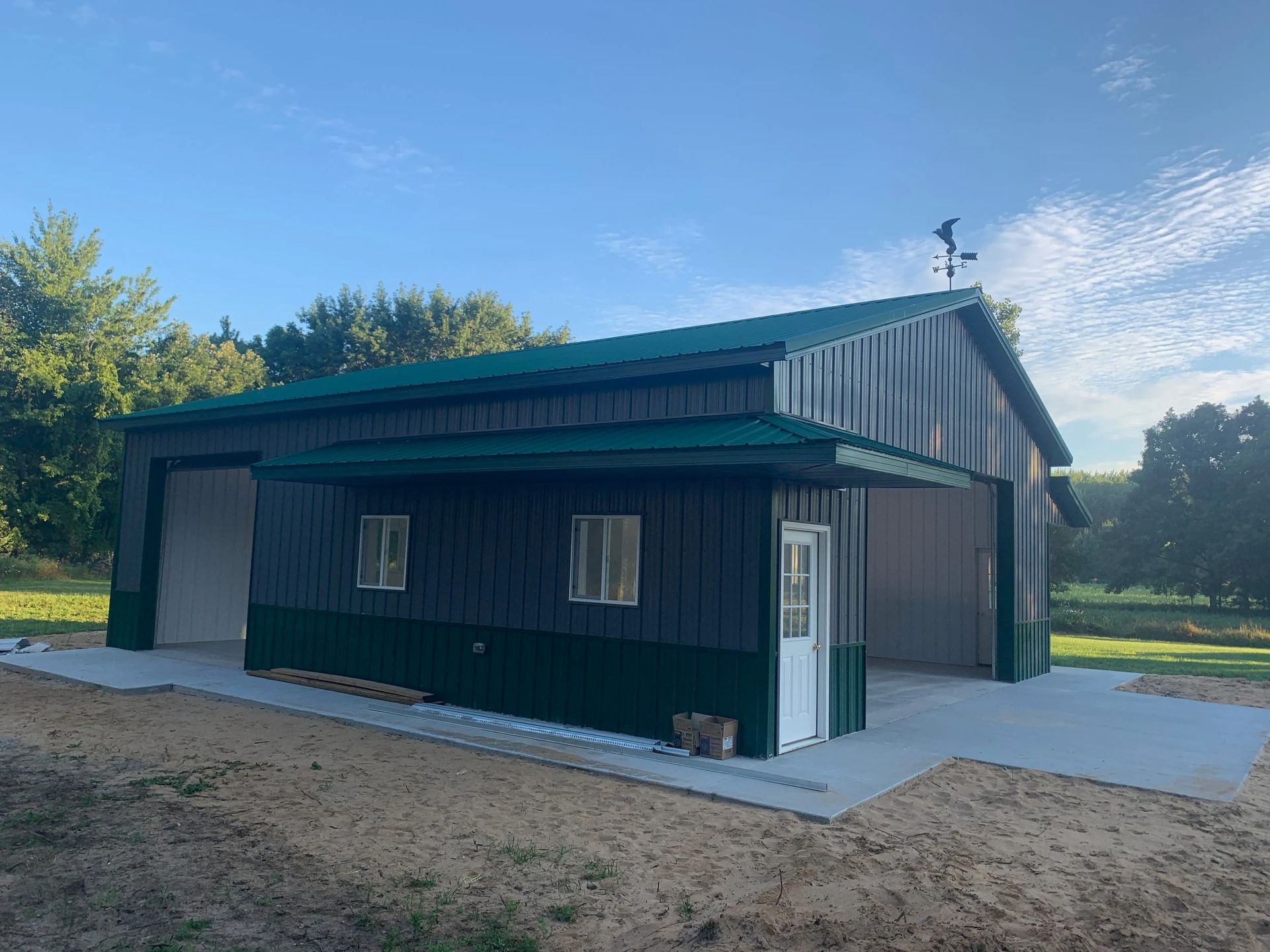 Small dark-green metal shed with a gray roof, white doors, and a concrete pad in a grassy yard.