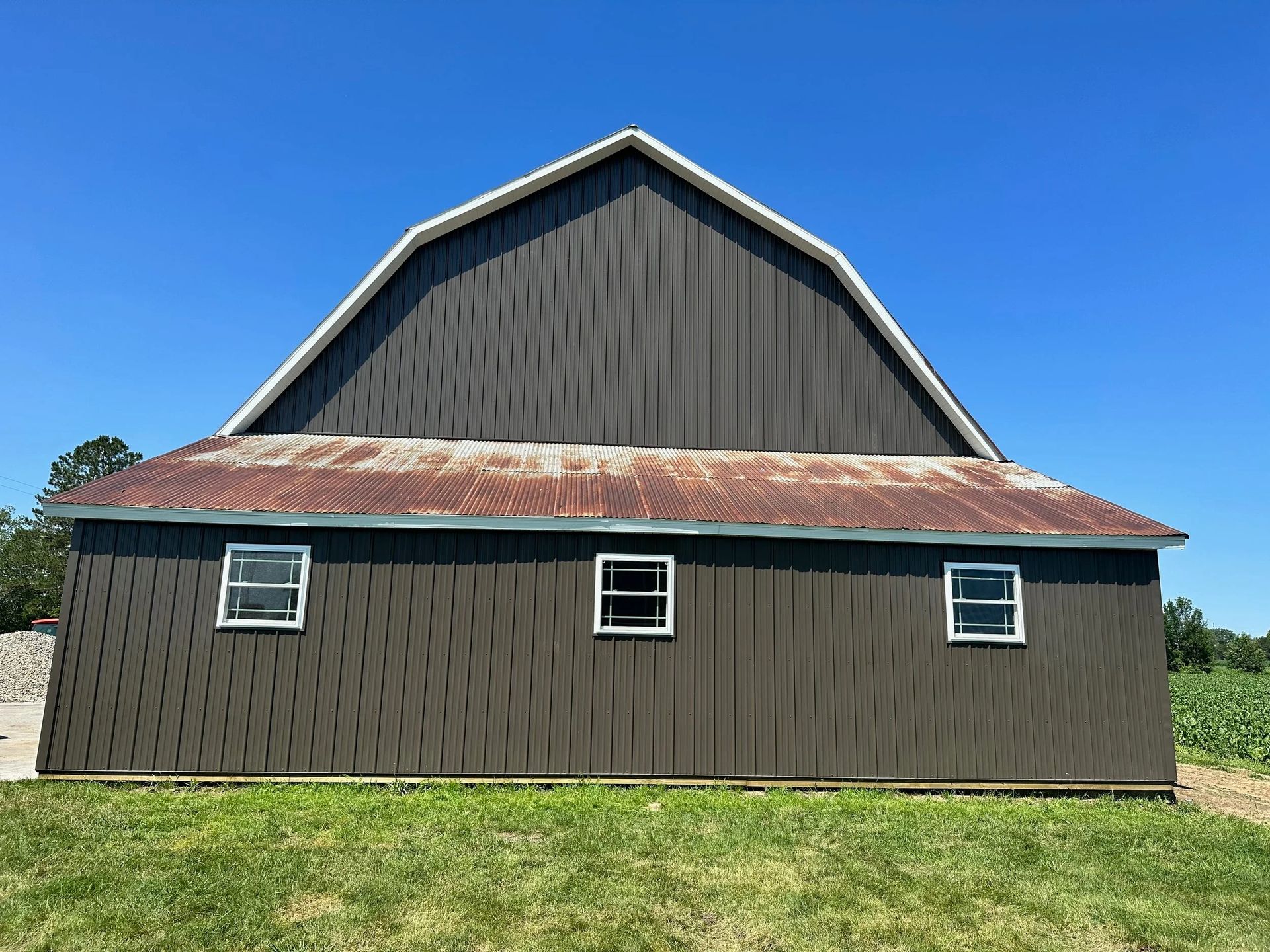 Dark barn with a red roof and white-trimmed windows on a grassy field under a blue sky