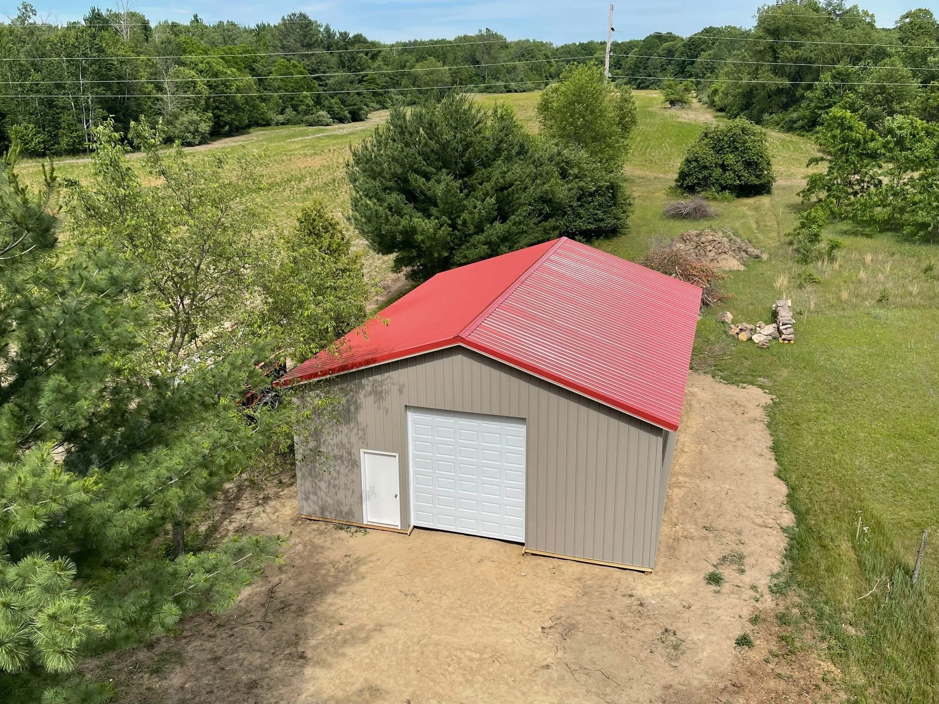 Small beige shed with a red metal roof in a grassy field, surrounded by trees and shrubs.