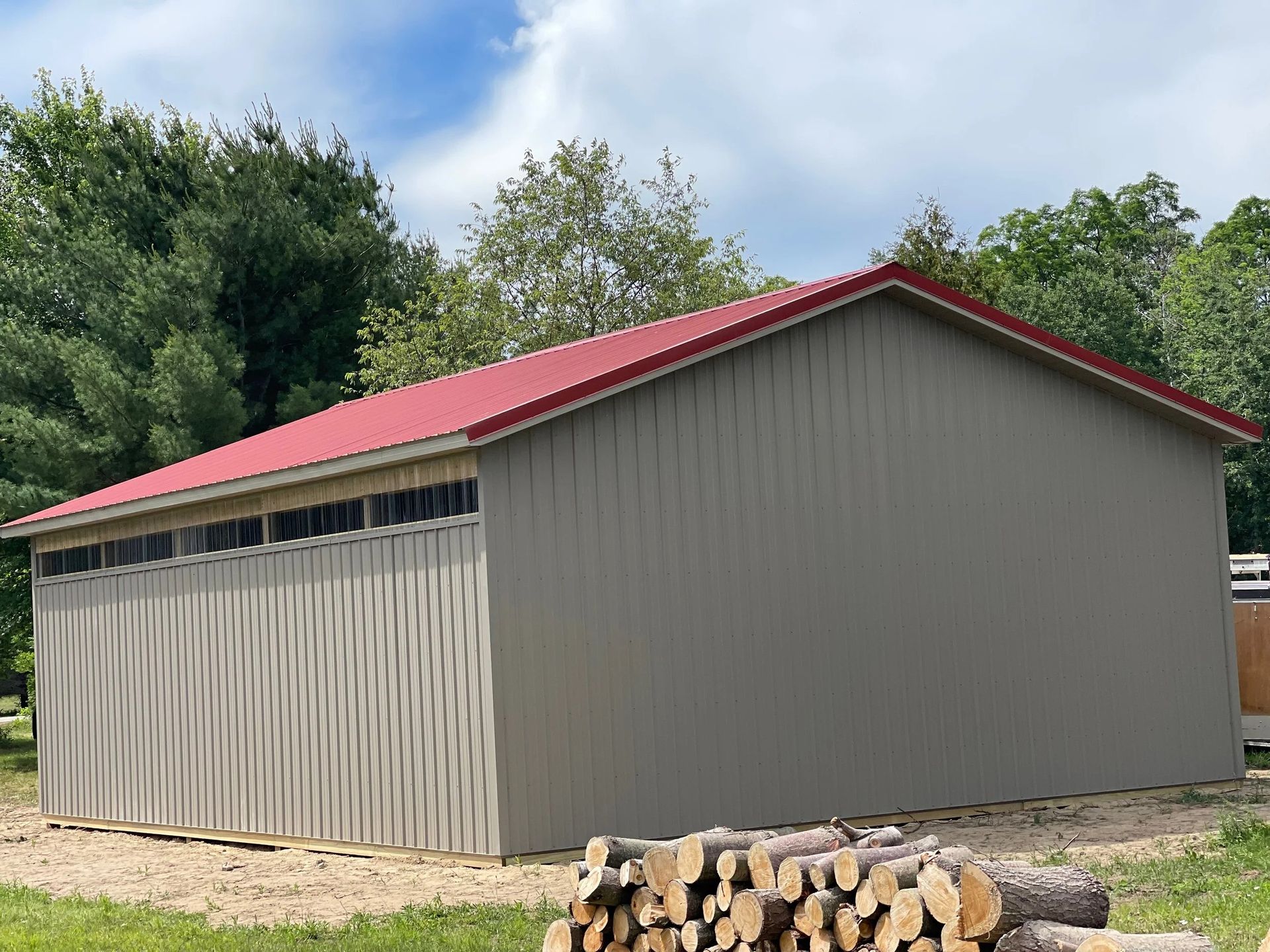 Gray shed with a red metal roof, roll-up door, and stacked firewood beside it in a grassy yard.