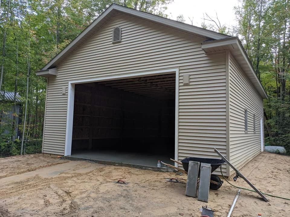 Detached beige garage with open door on a dirt lot, surrounded by trees