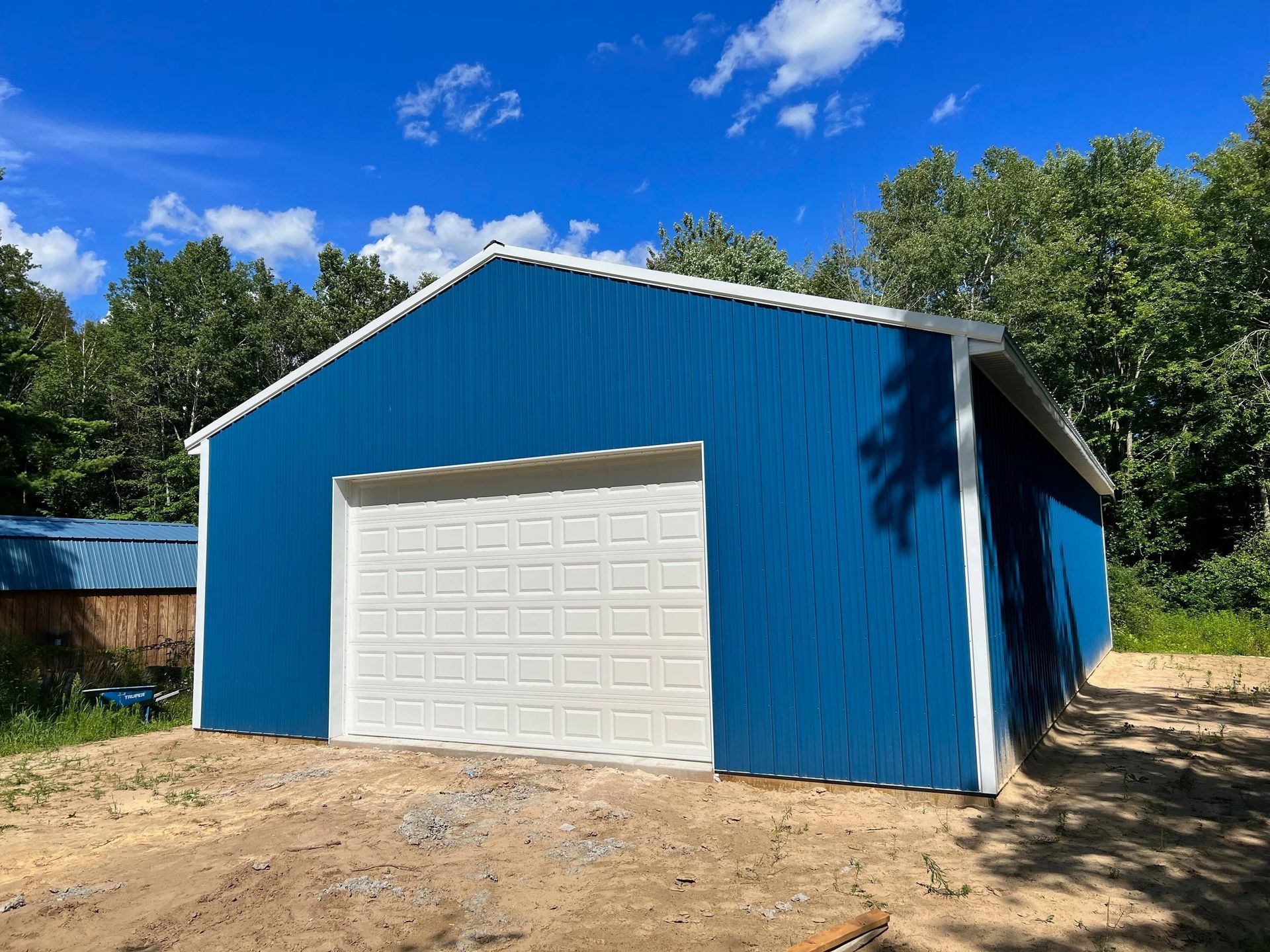 Blue garage with white door in a sunny wooded yard under a blue sky.