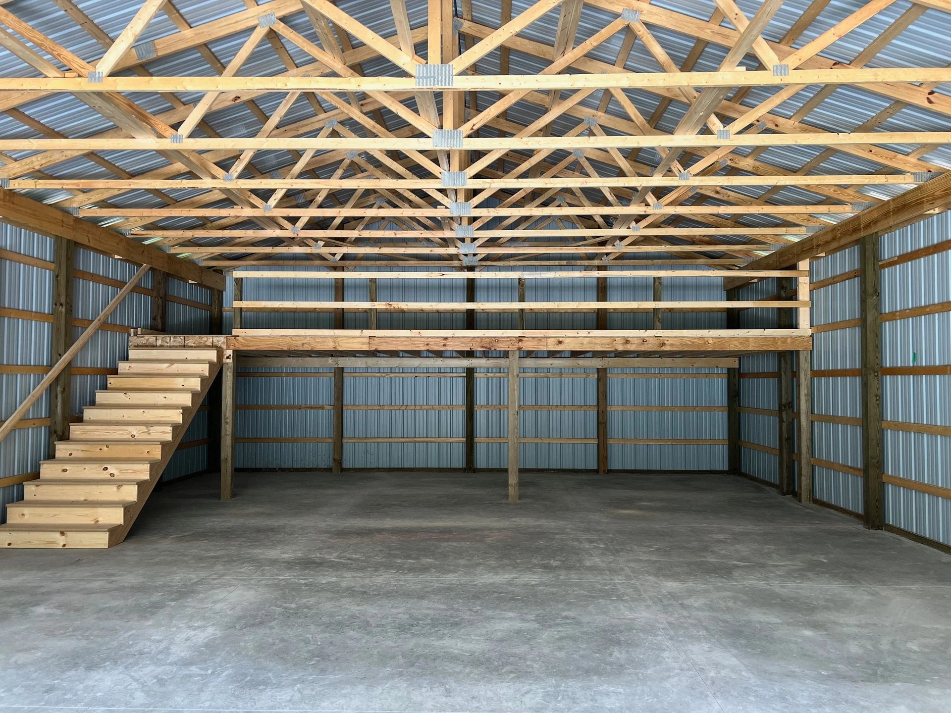 Empty wooden garage with loft, stairs, exposed roof trusses, and concrete floor