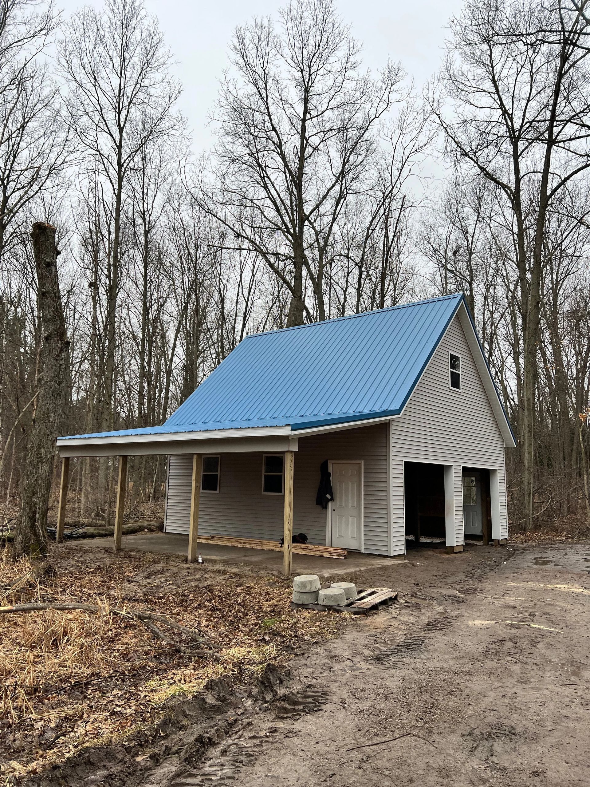 Small unfinished cabin with blue metal roof in a leafless forest, beside a dirt path.