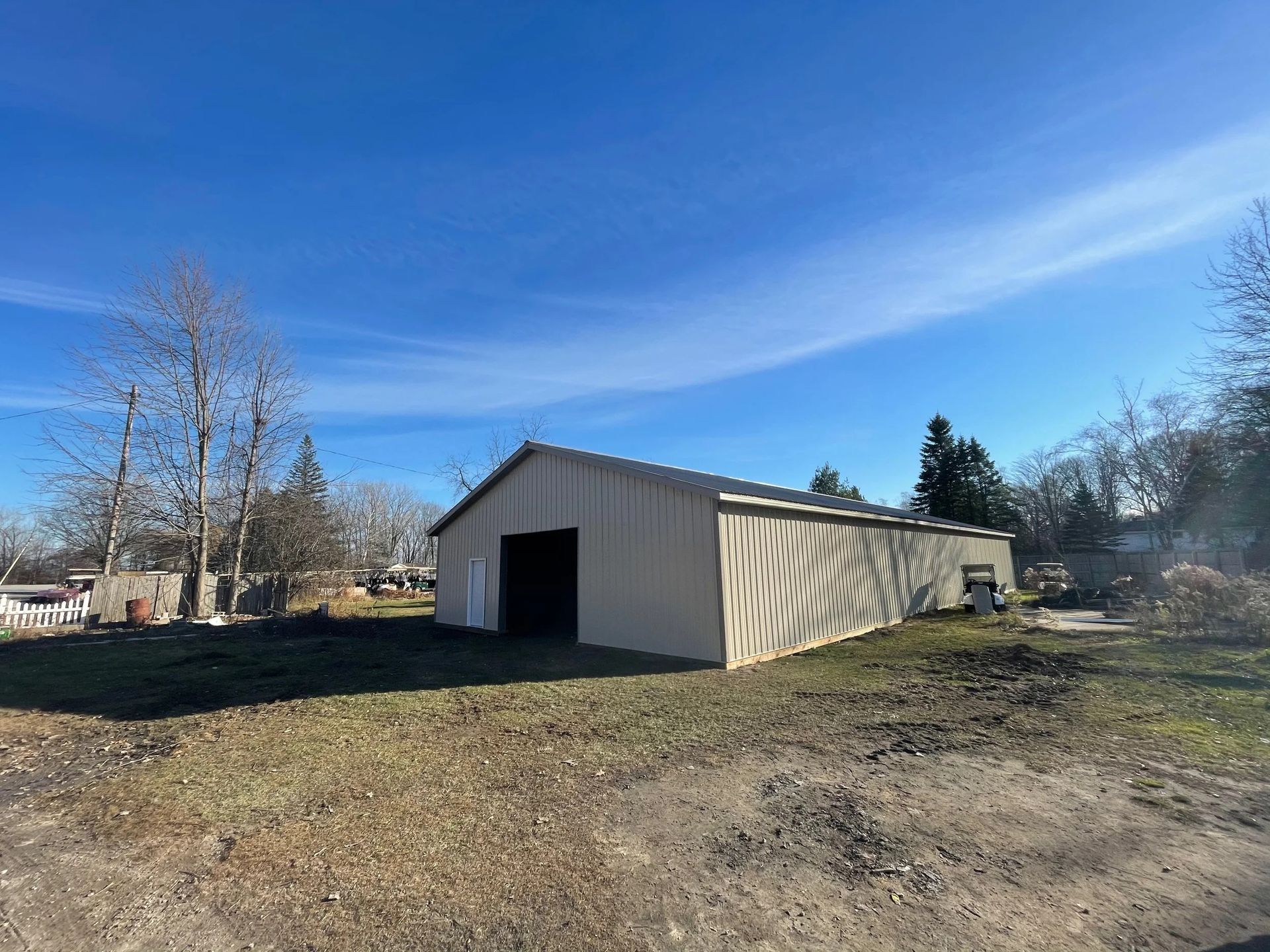 Large beige barn in a rural yard under a blue sky with bare trees and dirt foreground