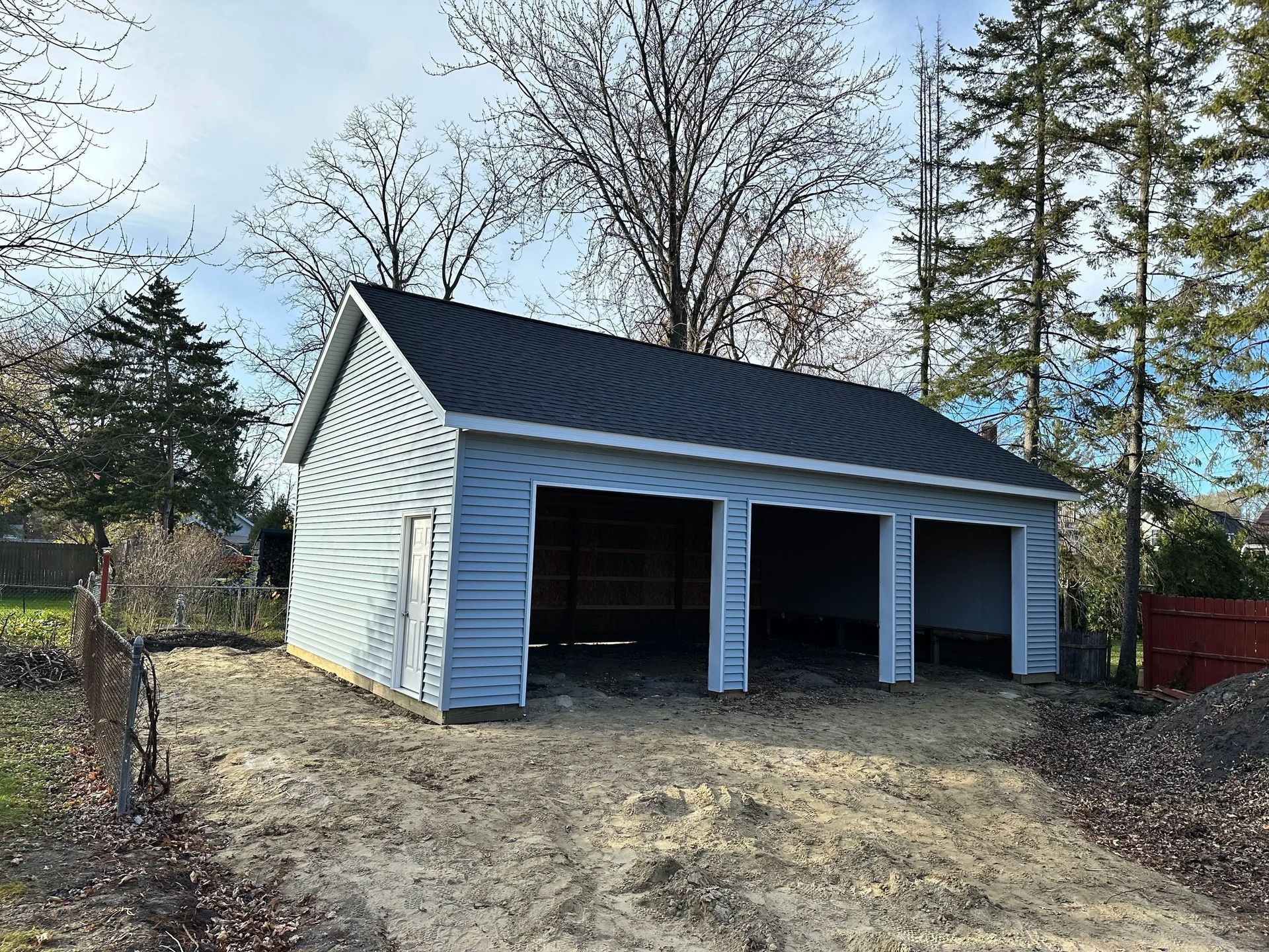 White garage with three open bays and a dark roof on a dirt lot, surrounded by bare trees