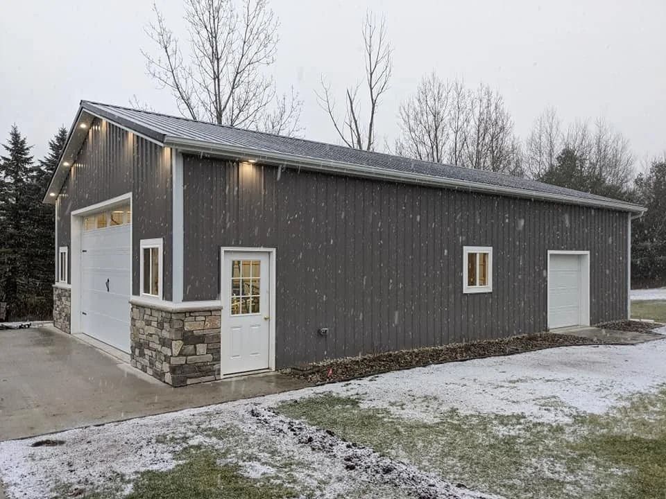 Gray metal barn-style building with stone accents and garage doors in a snowy rural setting