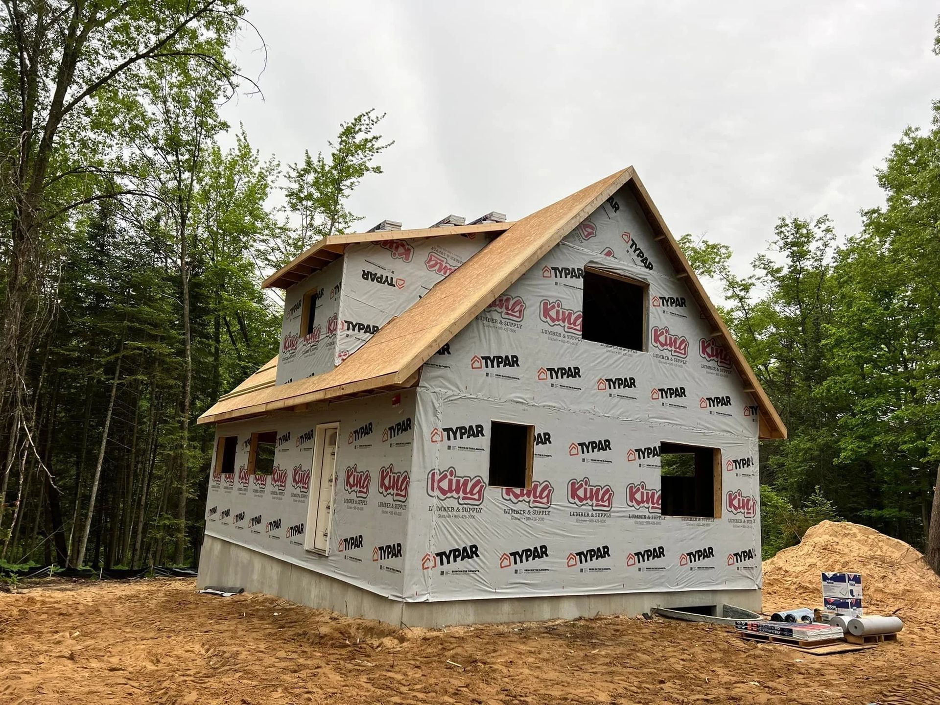 House under construction wrapped in housewrap, with tan roof sheathing and forested surroundings