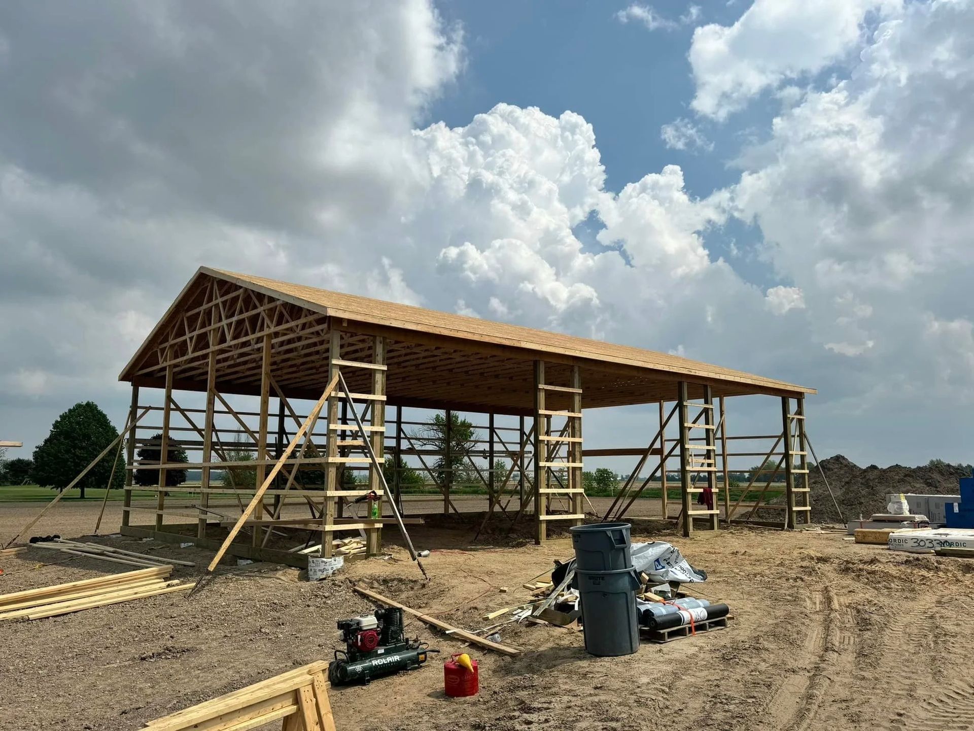 Wood-framed barn under construction on a gravel lot, with scaffolding and cloudy sky.