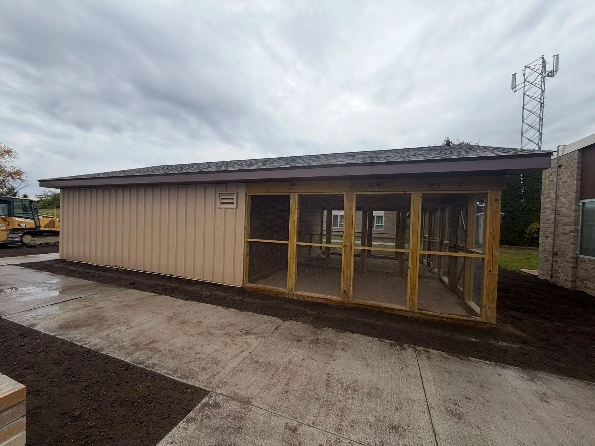 Small beige modular building with glass entrance doors beside a wet concrete path under a cloudy sky