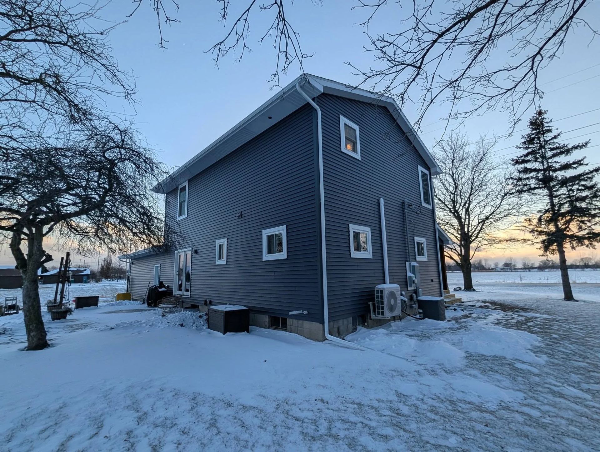 Snow-covered blue house at dusk with bare trees and a lake in the background