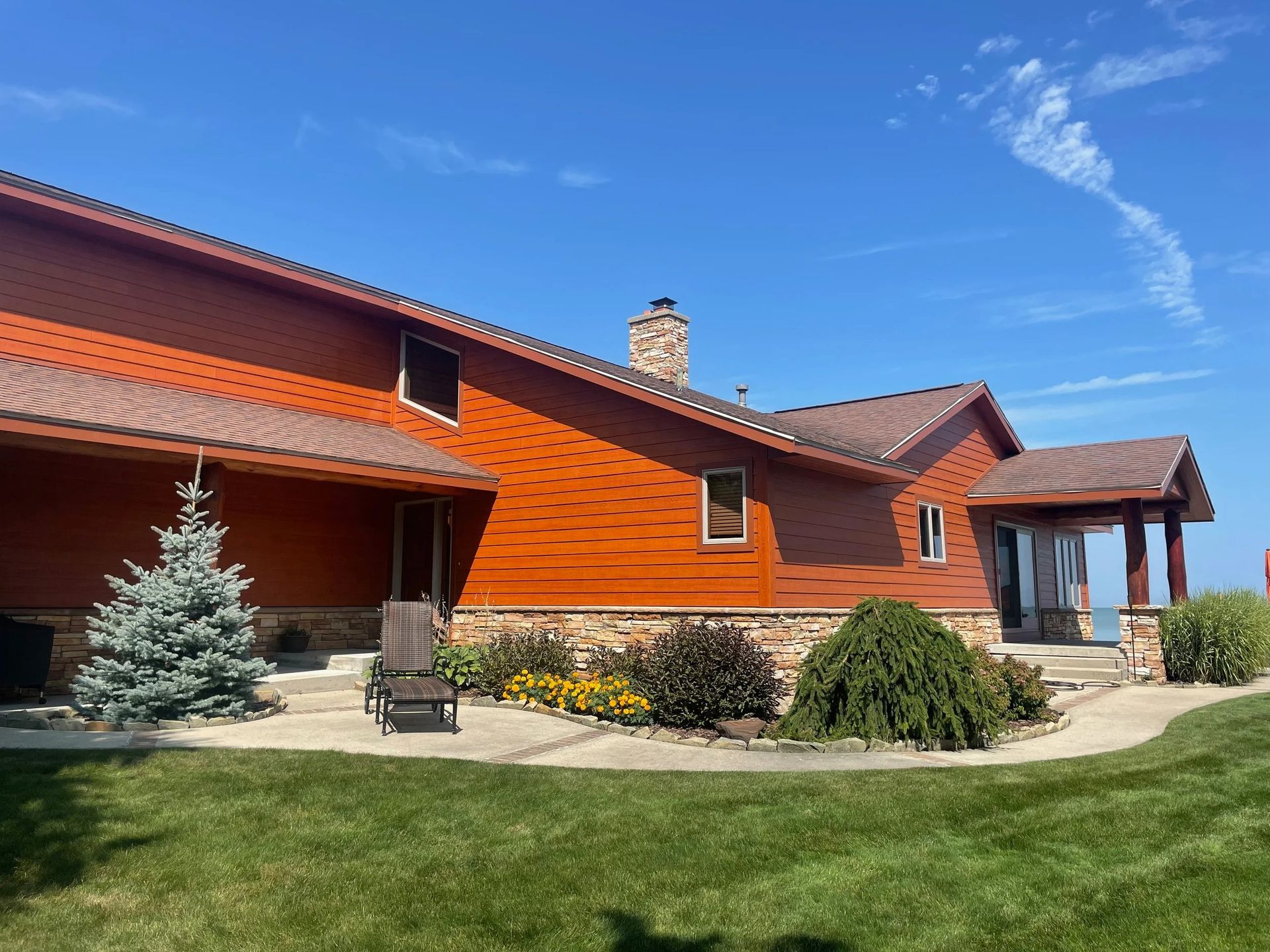 Red house with orange roof, stone chimney, patio chairs, and green lawn under a blue sky