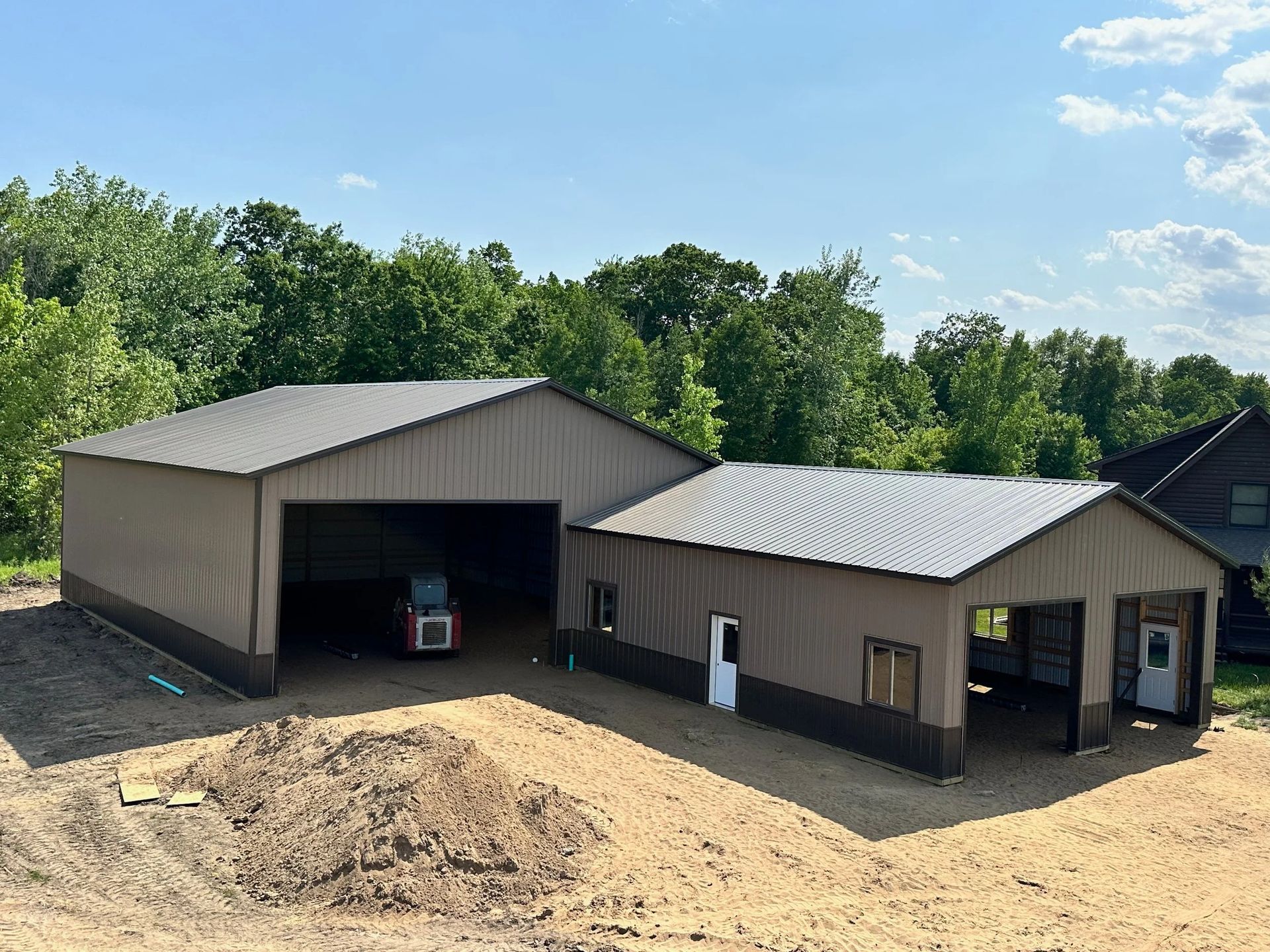 Two tan metal barns with open bays beside a dirt mound under a blue sky.