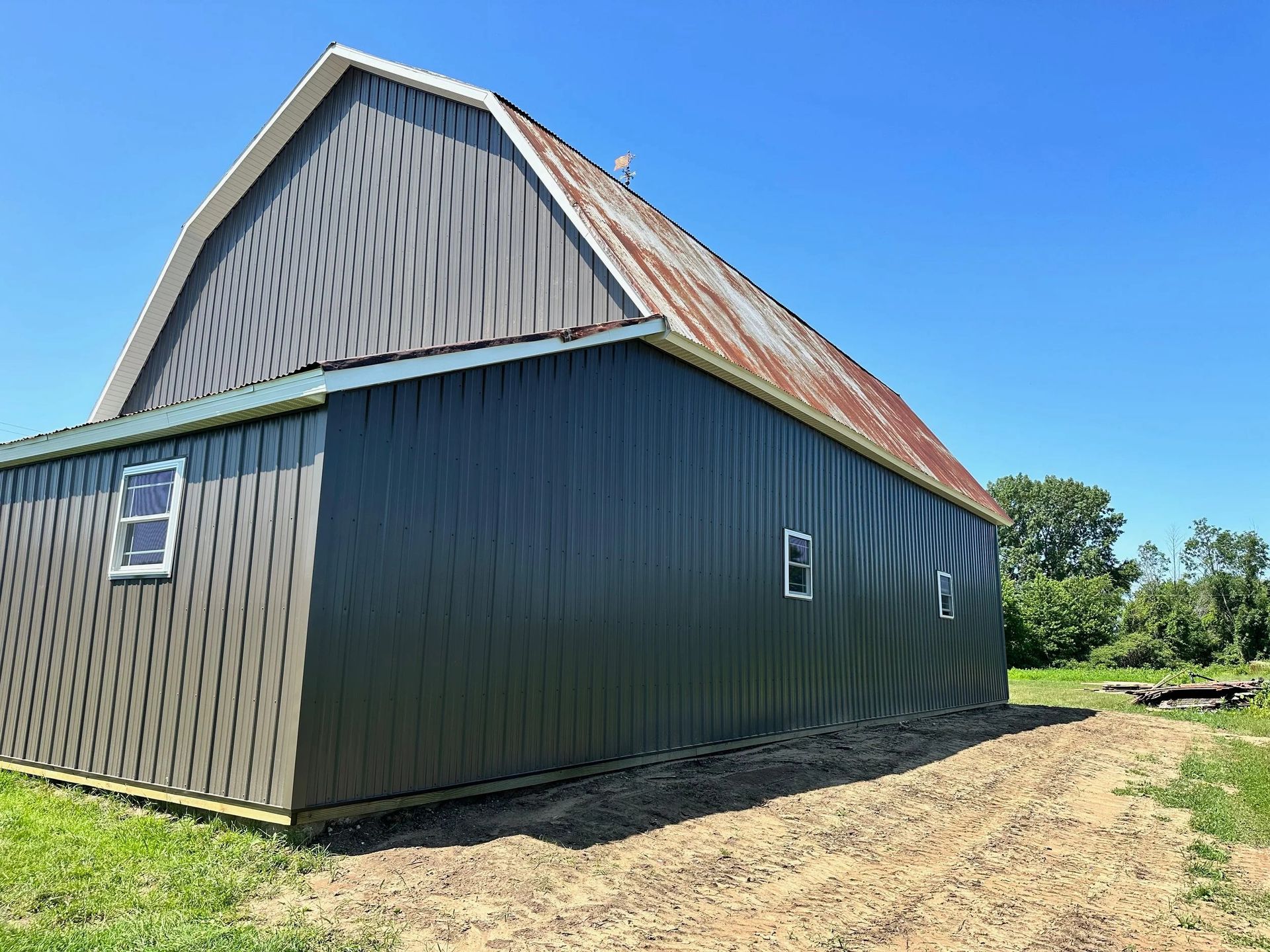 Large barn with dark siding and a rusted roof on a dirt lot under a blue sky