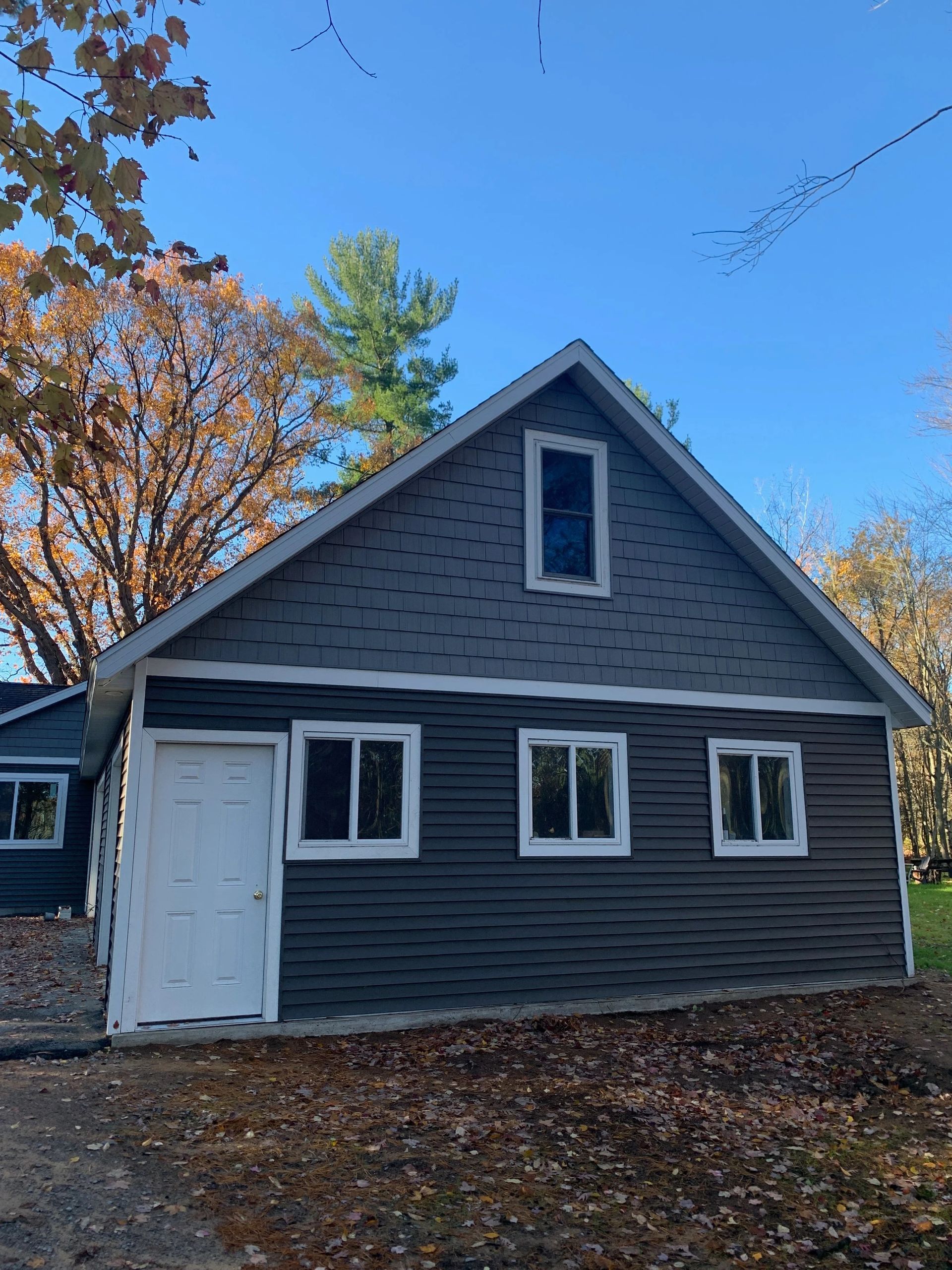 Gray house with white trim and a white door, set among autumn trees under a blue sky