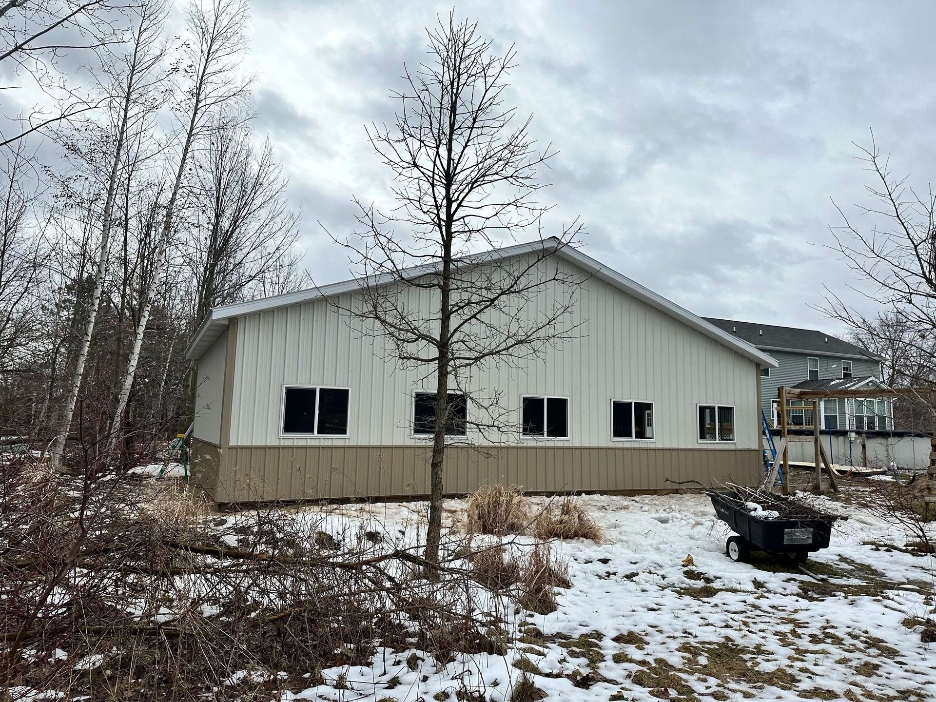 Snow-covered yard with a white metal building and bare trees under a cloudy sky