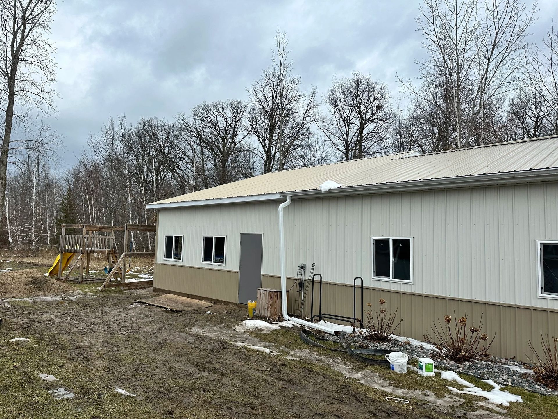 White metal building with gray roof in a muddy, leafless yard under overcast sky