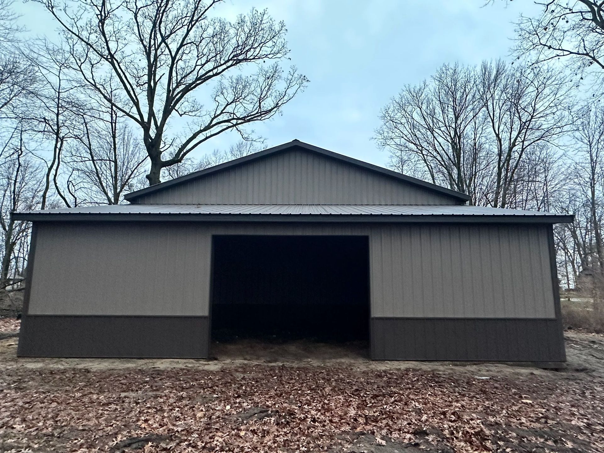 Gray barn with open doorway in a leaf-covered yard, framed by bare trees under an overcast sky