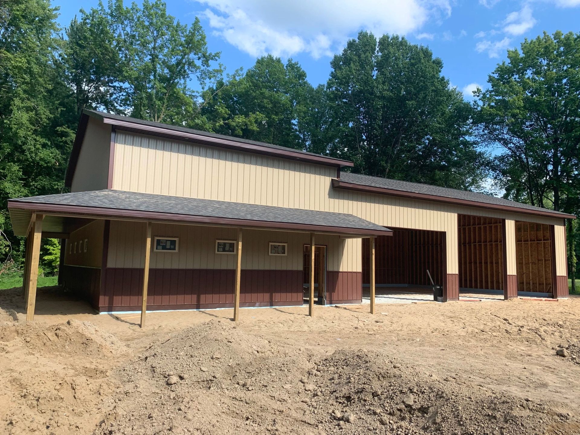 Rustic tan metal building with a covered porch on a dirt lot, surrounded by trees and blue sky.