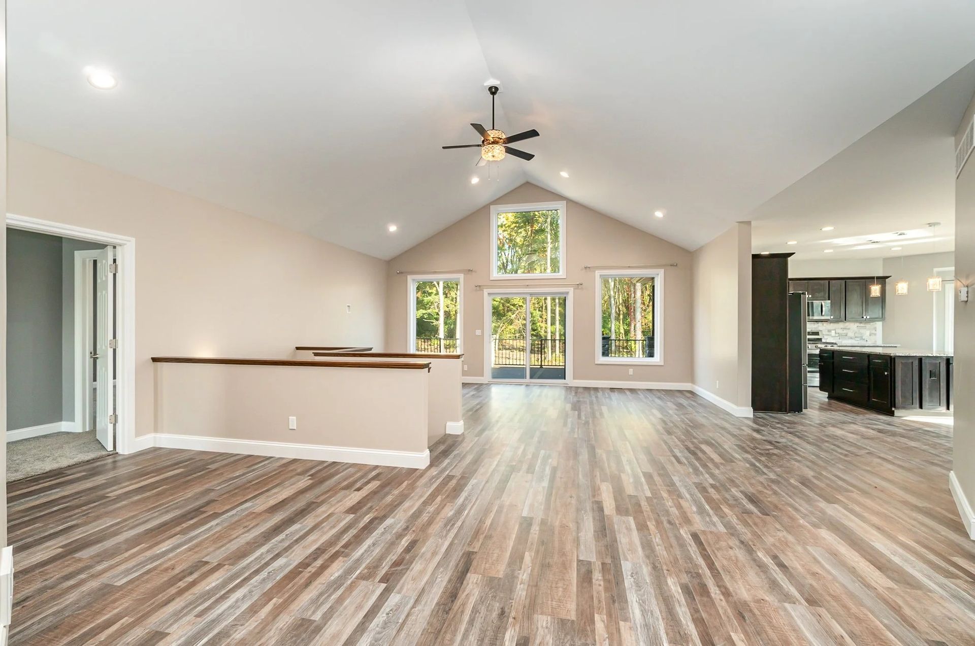 Spacious open-concept living room with vaulted ceiling, wood-look floors, and large windows overlooking greenery