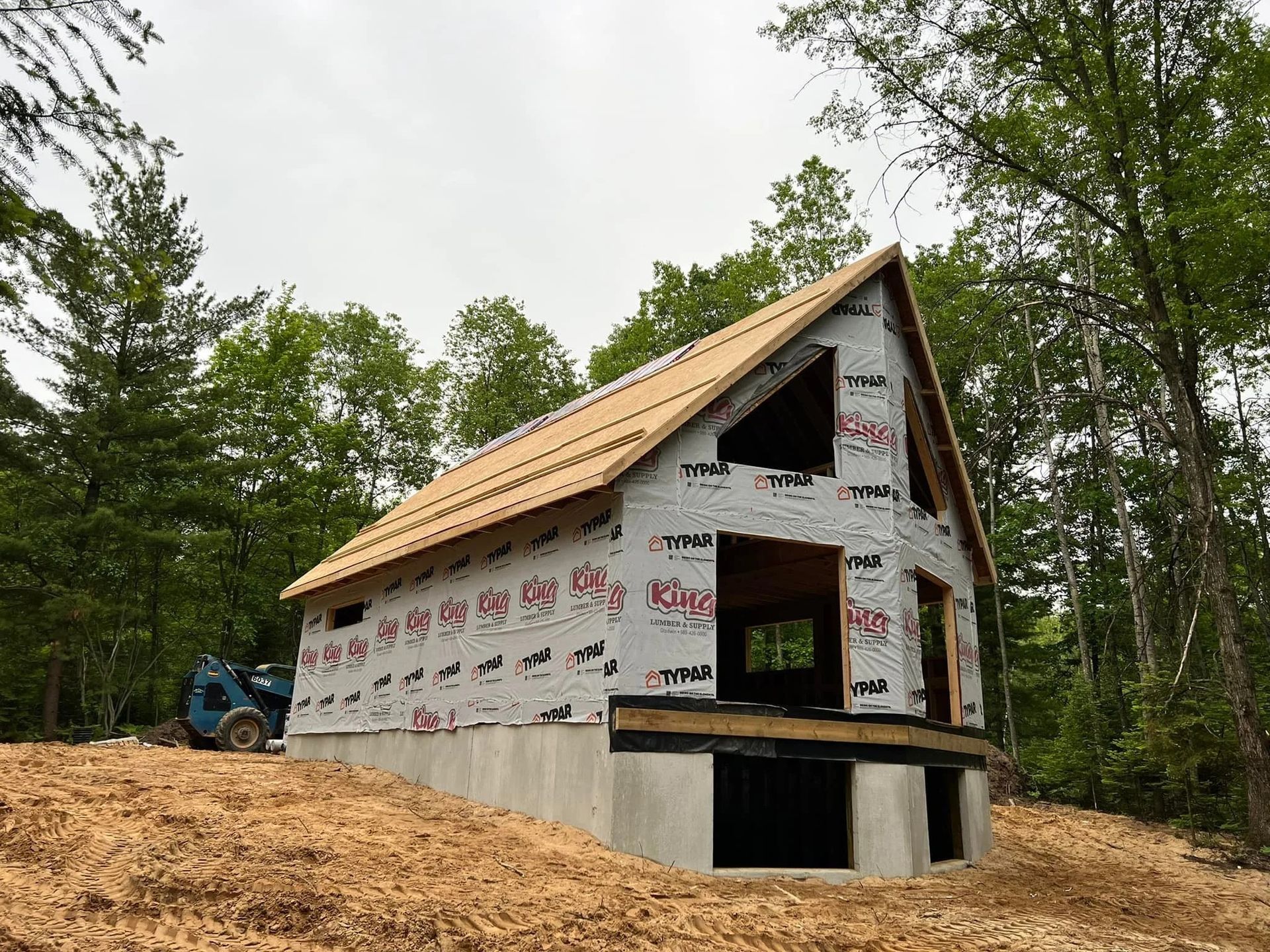 Partially built cabin with wrapped exterior and exposed roof framing in a wooded area