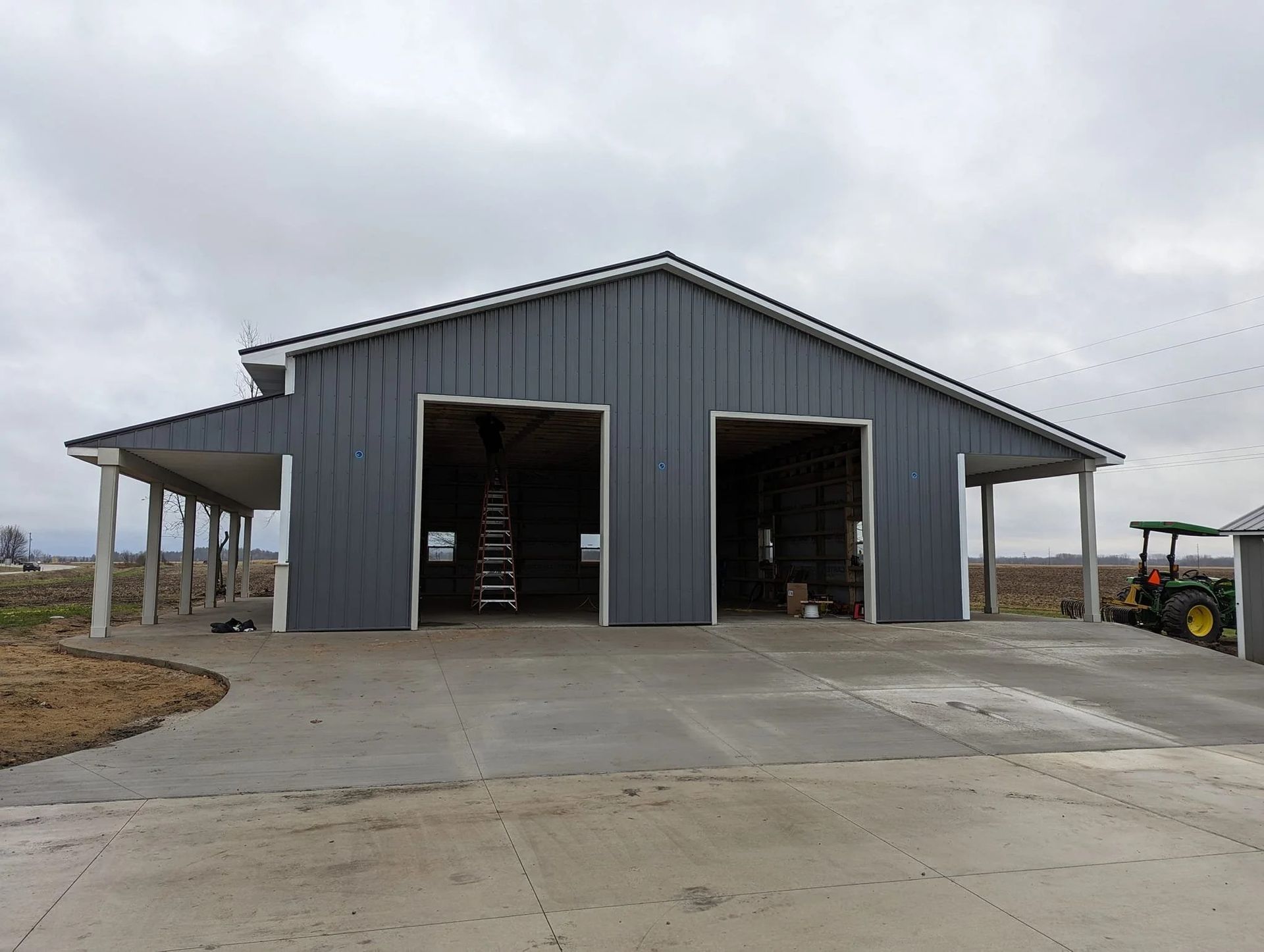 Gray metal barn with two open bays and a concrete driveway under a cloudy sky