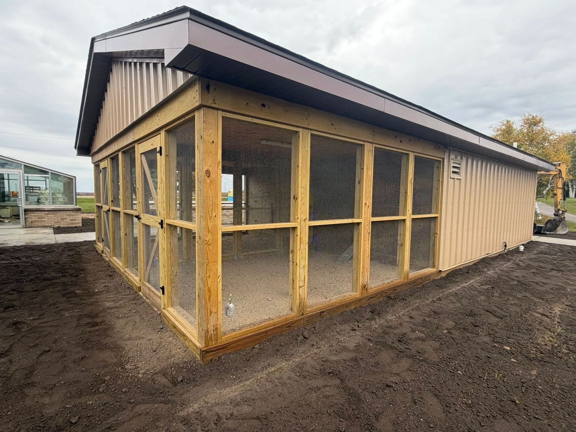 Wooden outdoor aviary with wire mesh panels and a sloped dark roof on muddy ground.