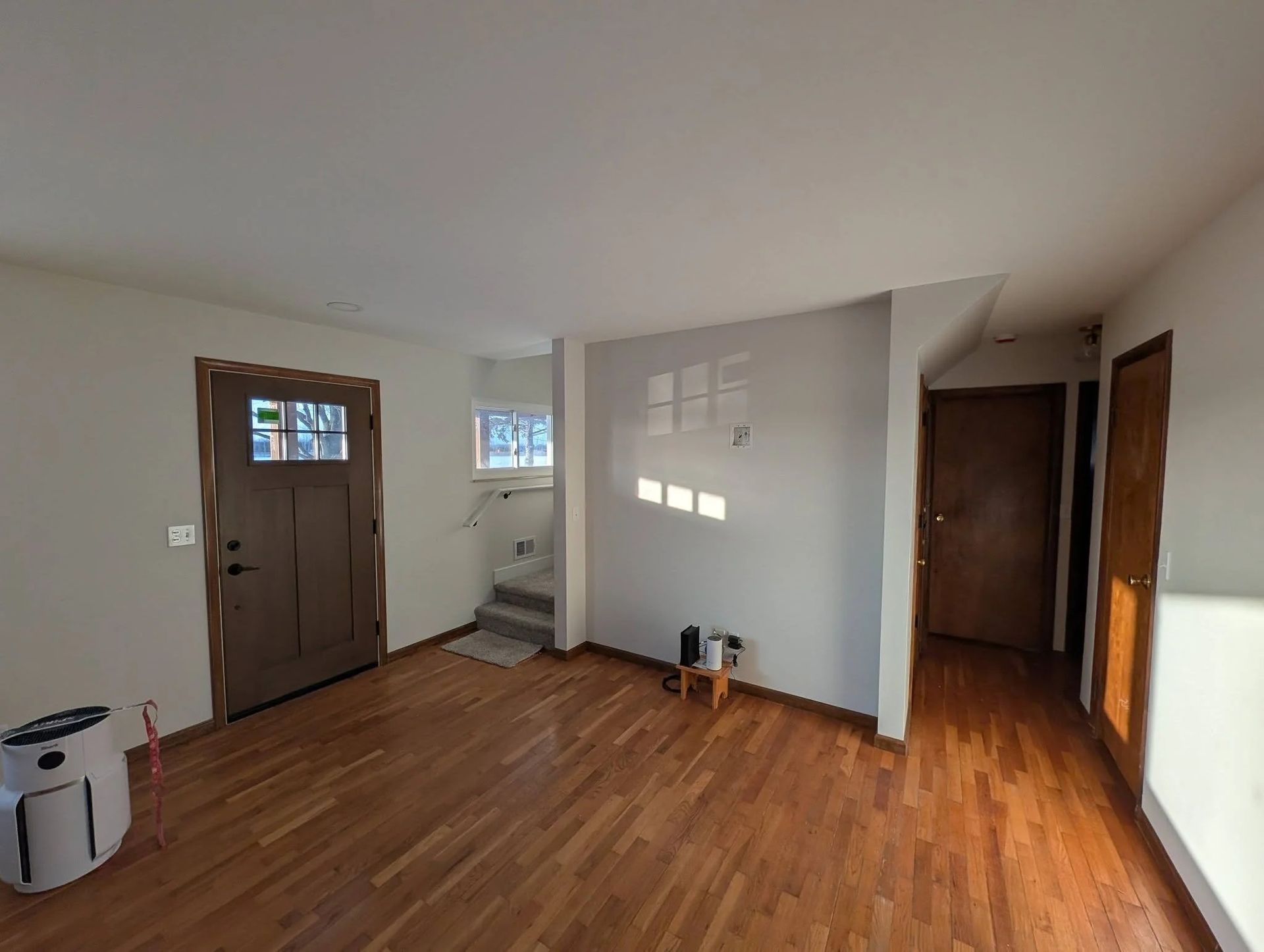 Empty living room with wood floors, front door, stairs, and sunlight on white walls