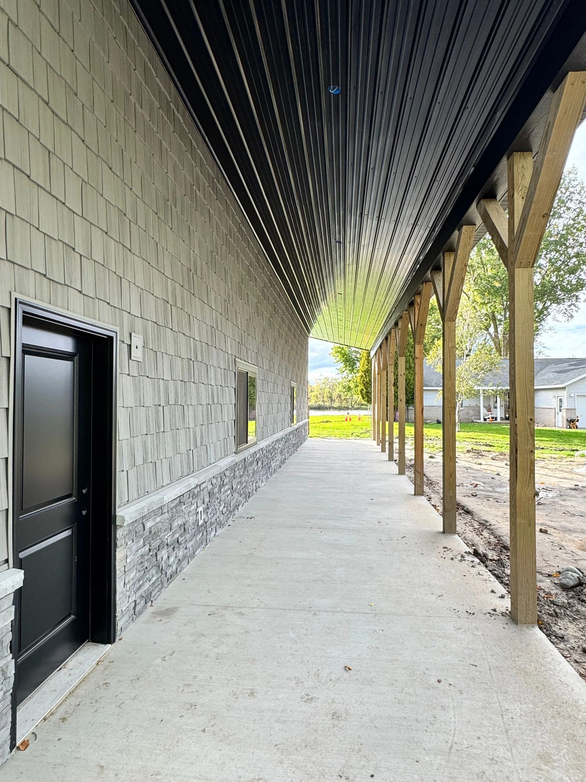 Covered stone walkway beside a black door, with wooden posts and a grassy view beyond.