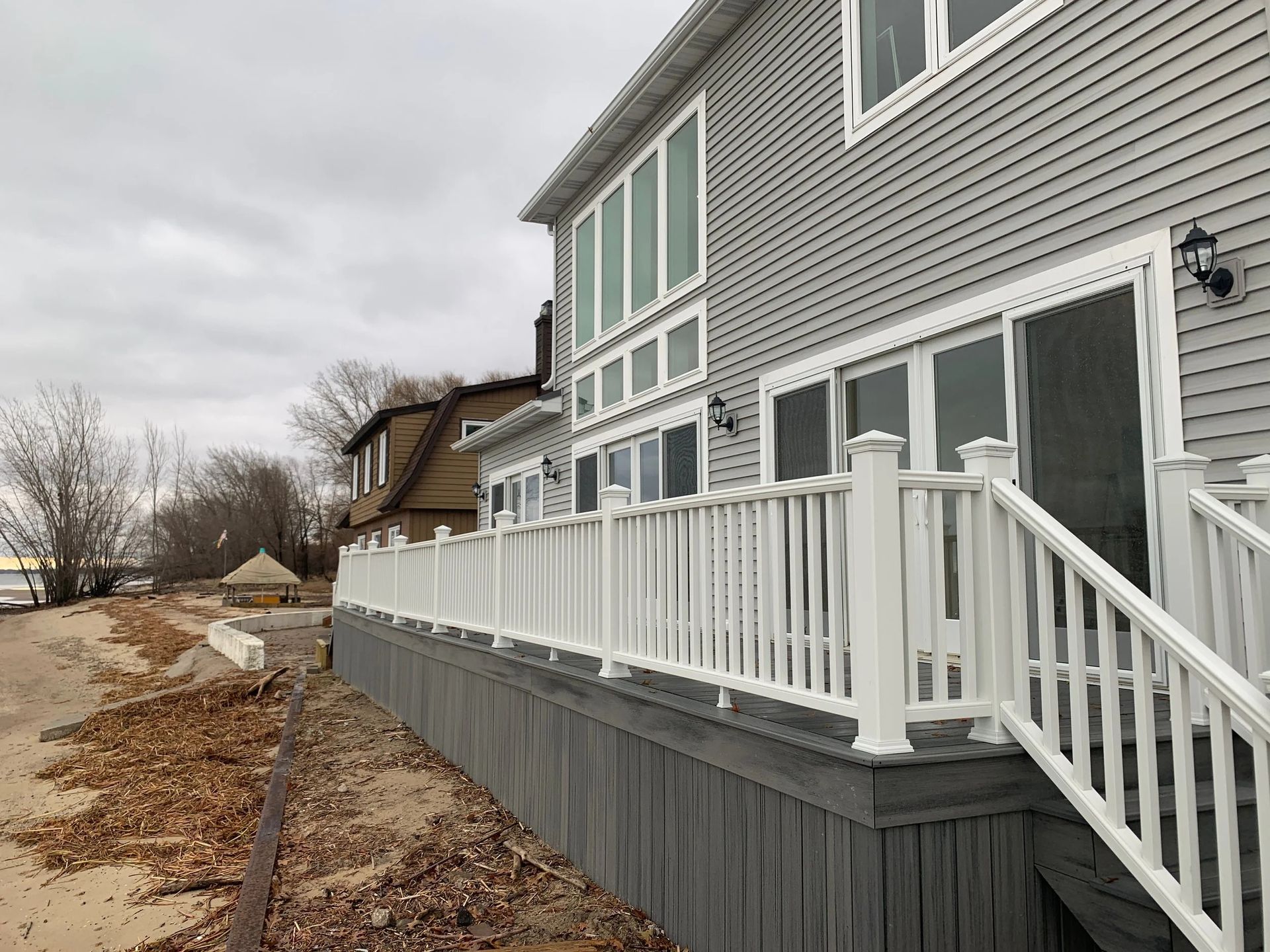 Gray house with white deck railings beside a rocky shoreline under an overcast sky