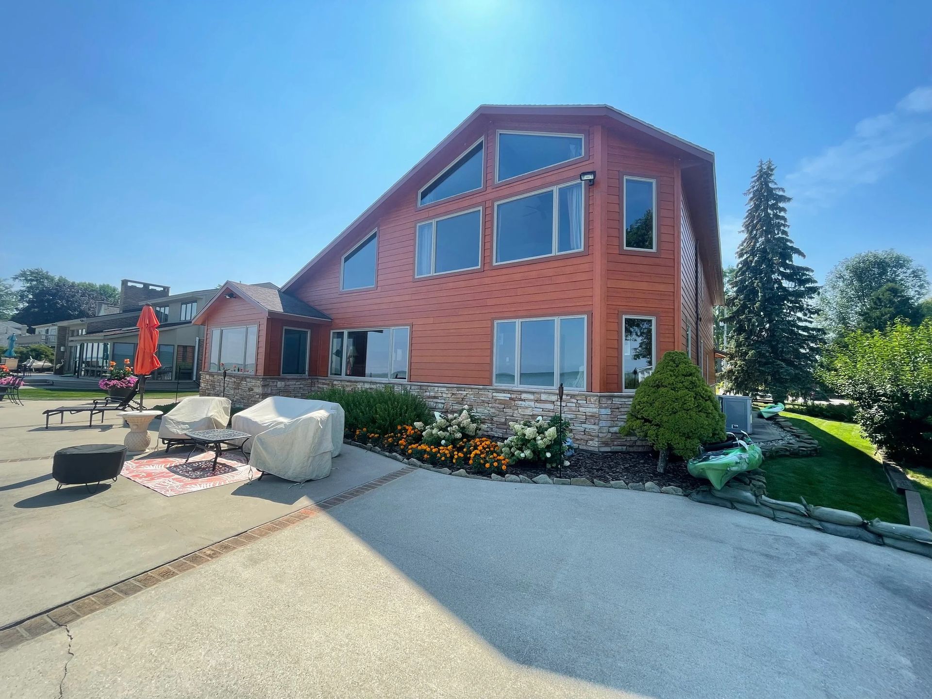 Red brick house with large windows, driveway, patio seating, and landscaped garden on a sunny day