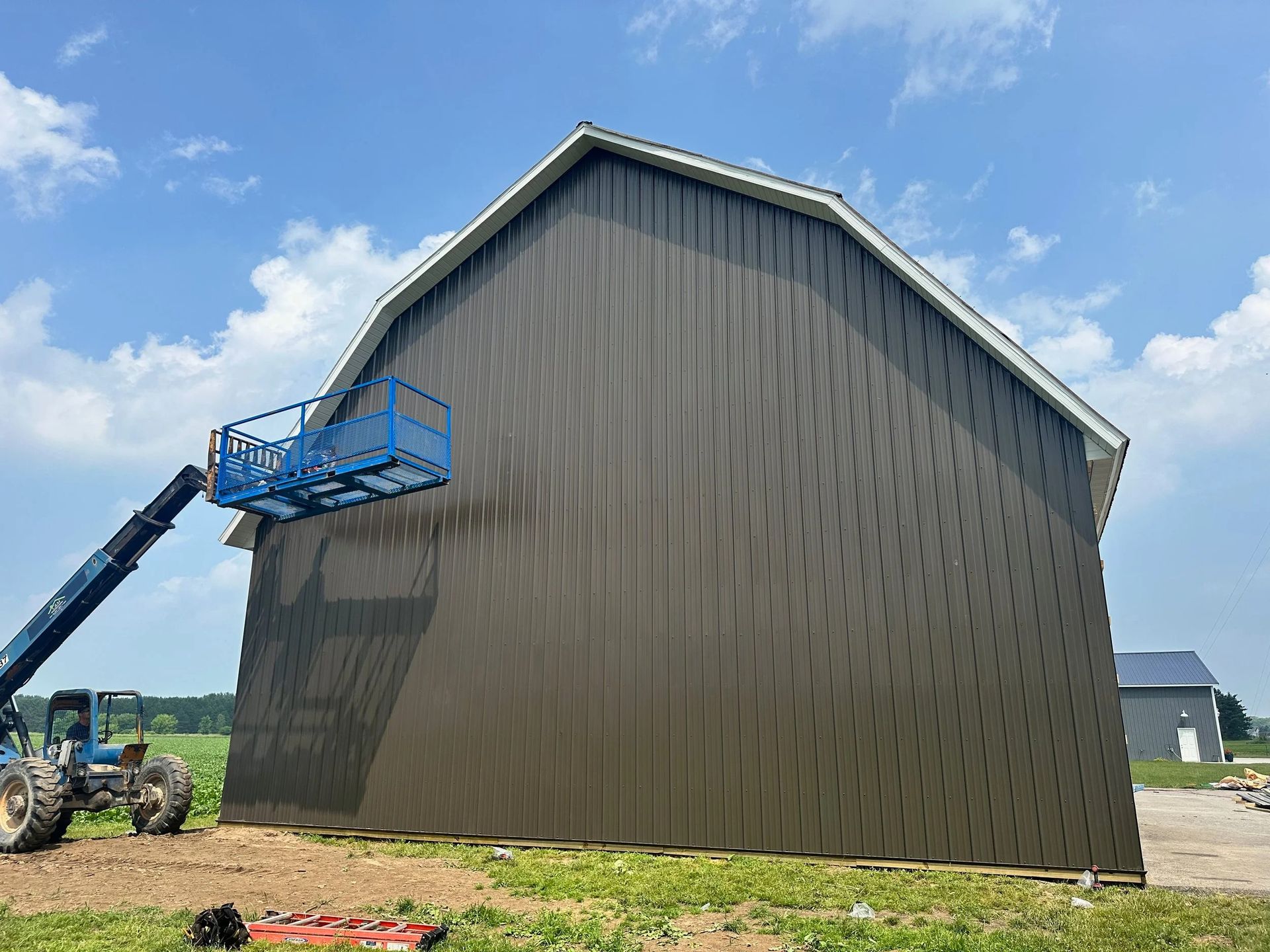 Blue cherry picker reaching up to a tall brown barn under a blue sky.