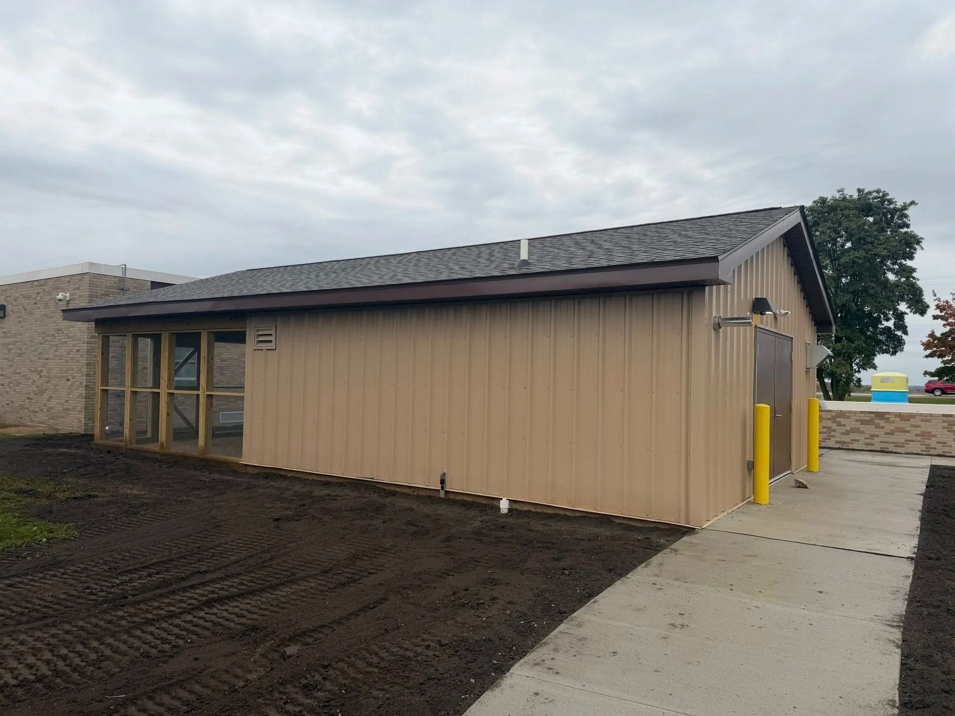 Single-story tan building with dark roof, windows, and concrete walkway under a cloudy sky.