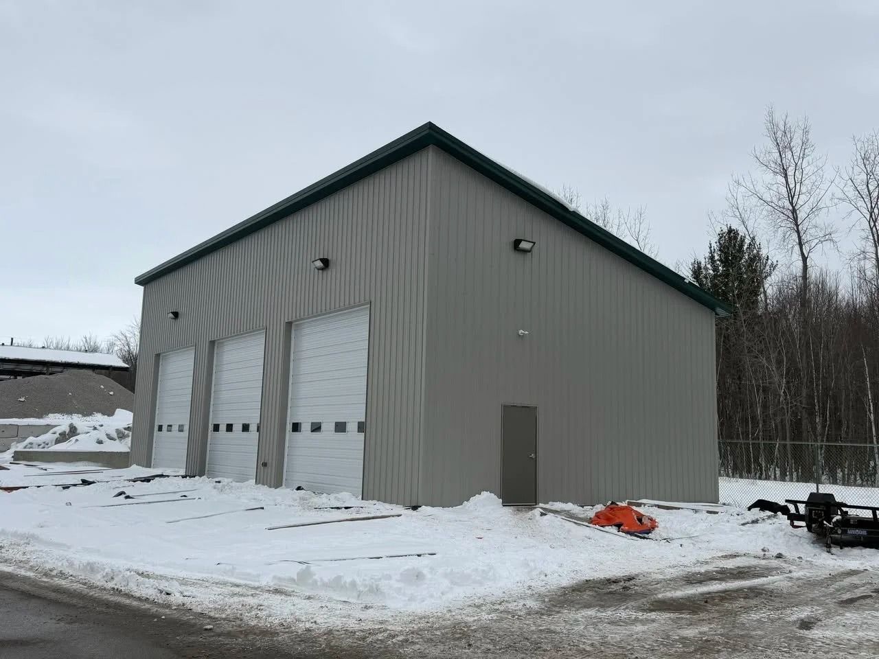 Snowy yard with a gray metal garage building and three white roll-up doors under an overcast sky